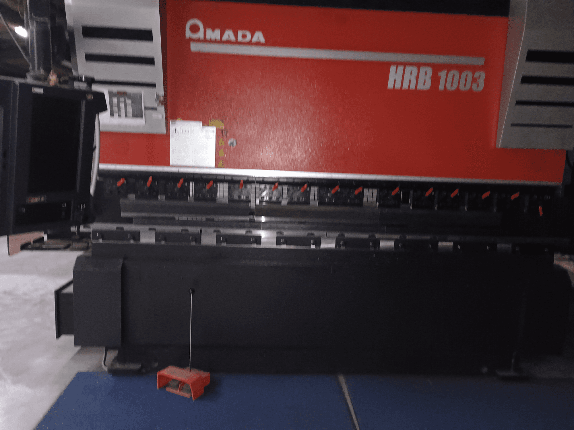 Amada HRB 1003 preses bremze, skats no priekšpuses; vadības panelis ar sarkanām pogām un melnu korpusu, ievietots rūpnieciskā vidē.