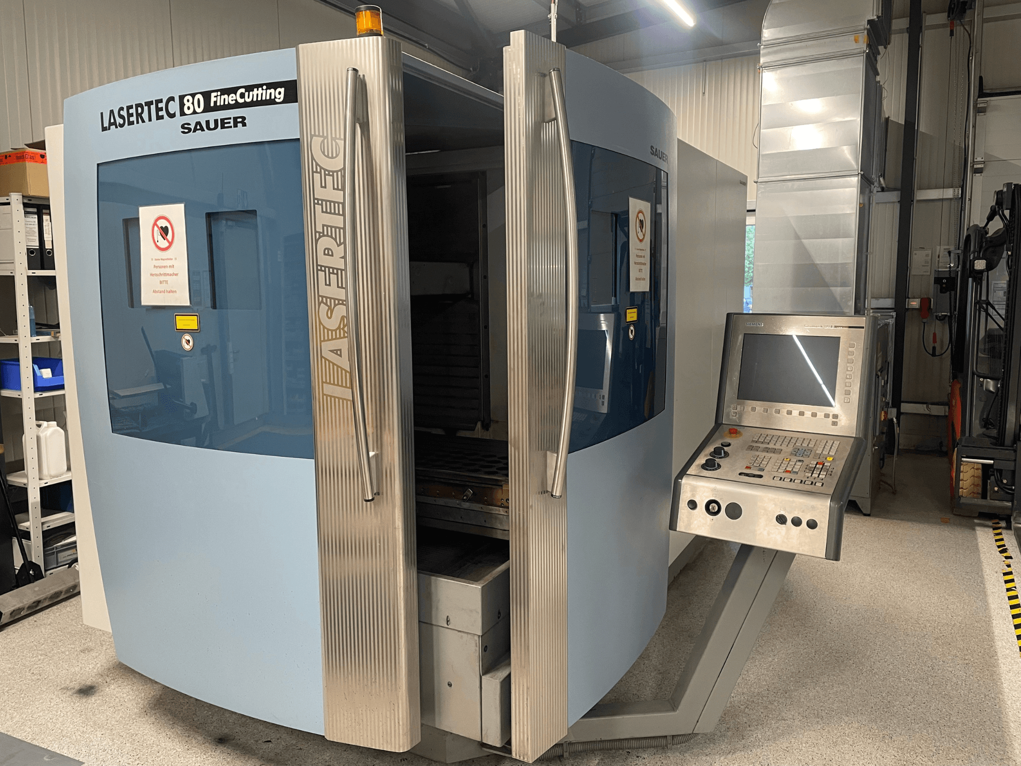 DMG Sauer Lasertec80 FineCutting metāla lokšņu apstrādes iekārtas priekšskats ar vadības paneli, atvērtām durvīm un redzamām drošības zīmēm.