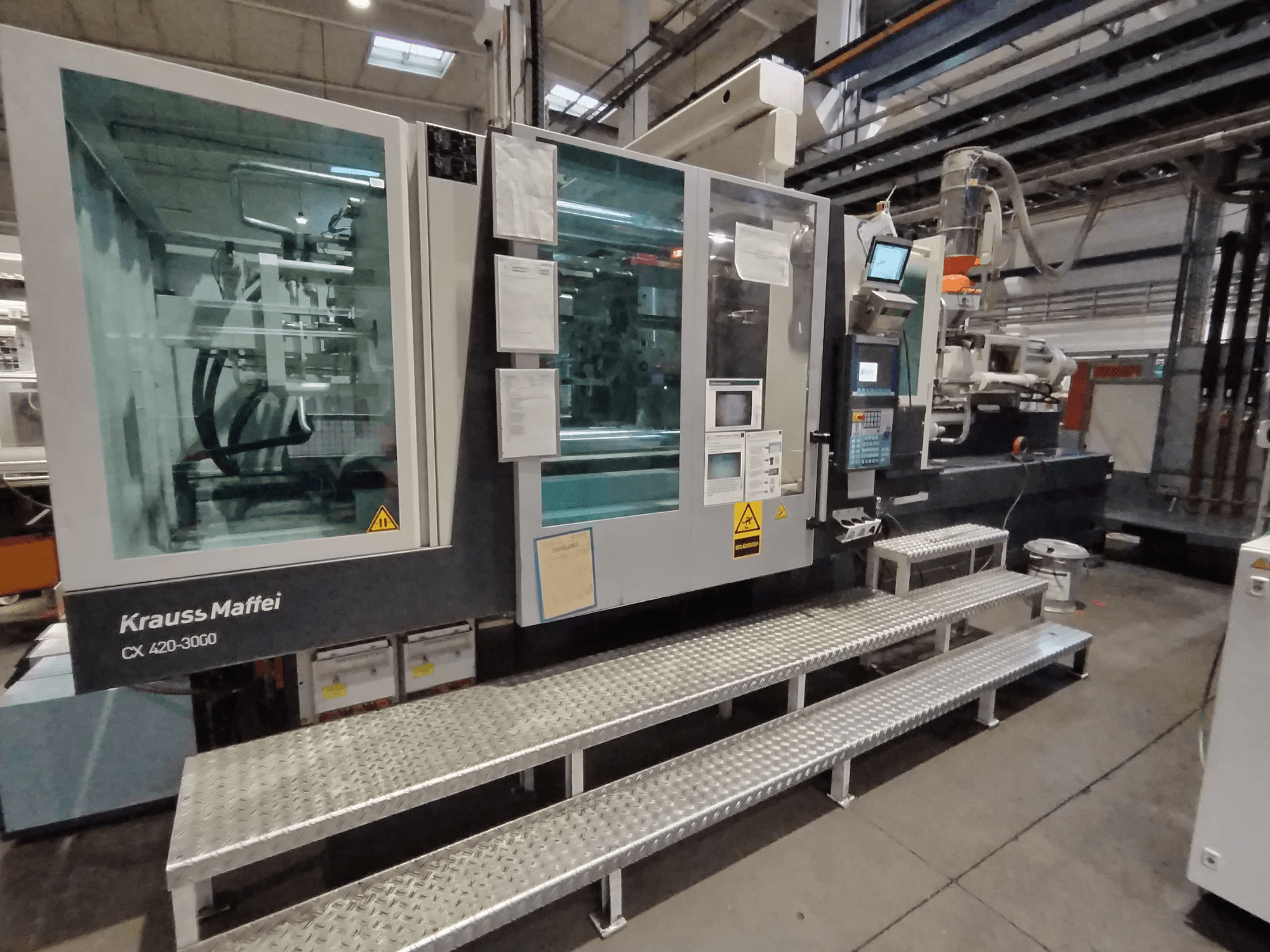 Krauss Maffei CX 420-3000 iesmidzināšanas veidošanas iekārta, skats no sāniem, ar redzamu vadības paneli un drošības zīmēm, novietota uz metāla pakāpieniem.