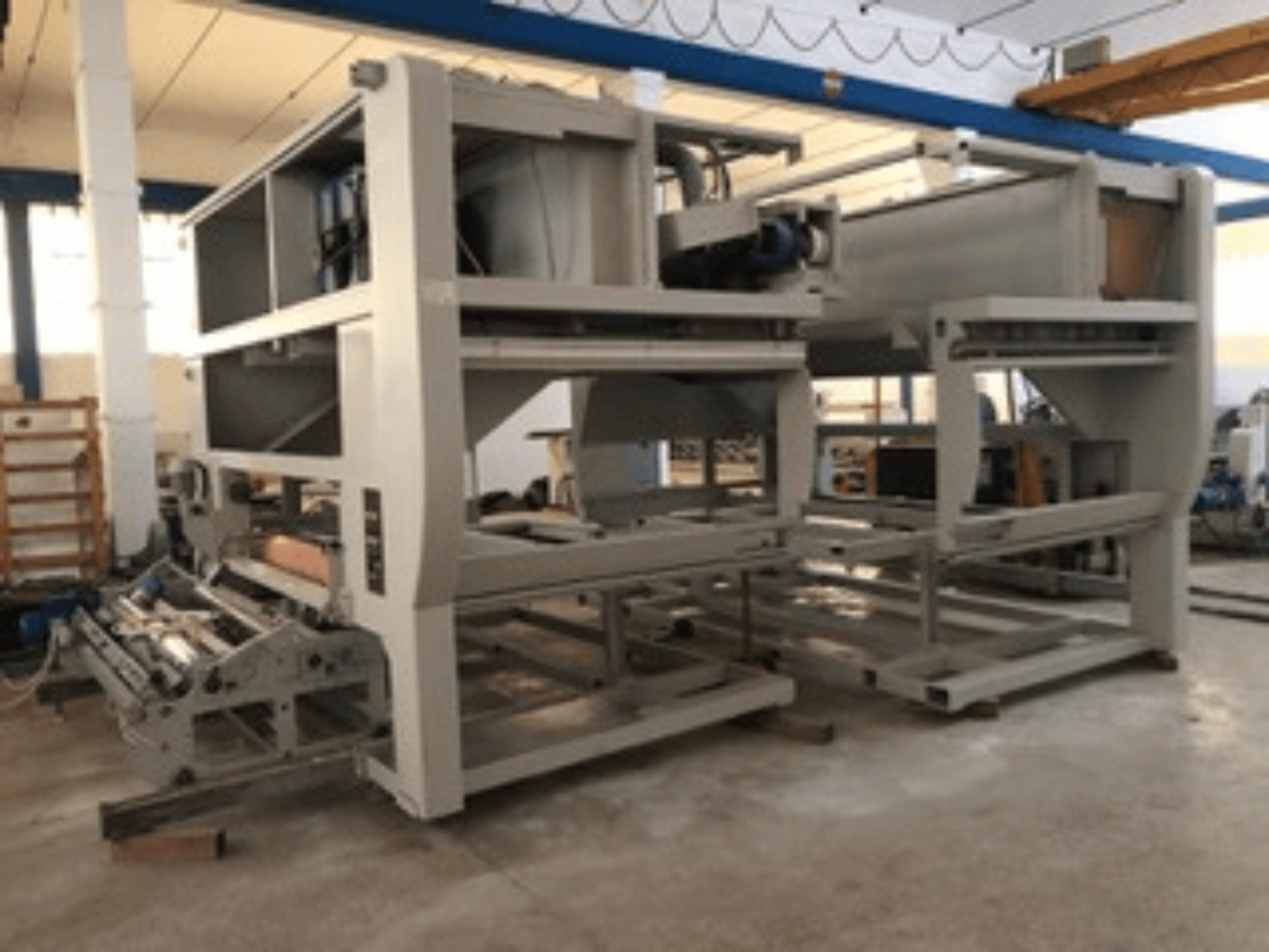 Rūpnieciskā mašīna ar vairākiem konveijera veltņiem un lielu konstrukciju; skats no sāniem, pelēkā krāsā, atrodas labi apgaismotā rūpnīcā.