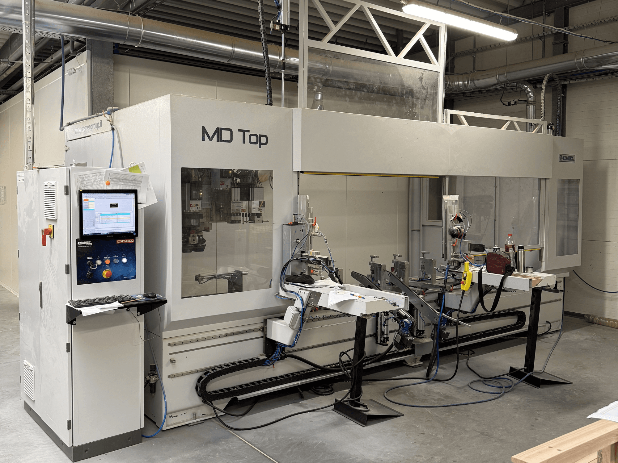 MD Top CNC mašīna, skatoties no priekšpuses, ar vadības paneli, robotizētām rokām un darba vietu ar instrumentiem un materiāliem.