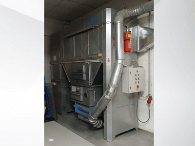 Rūpnieciskā putekļu savācēja iekārta, pelēks metāls, skats no priekšpuses; izplūdes kanāls, vadības panelis un ugunsdzēšamais aparāts.