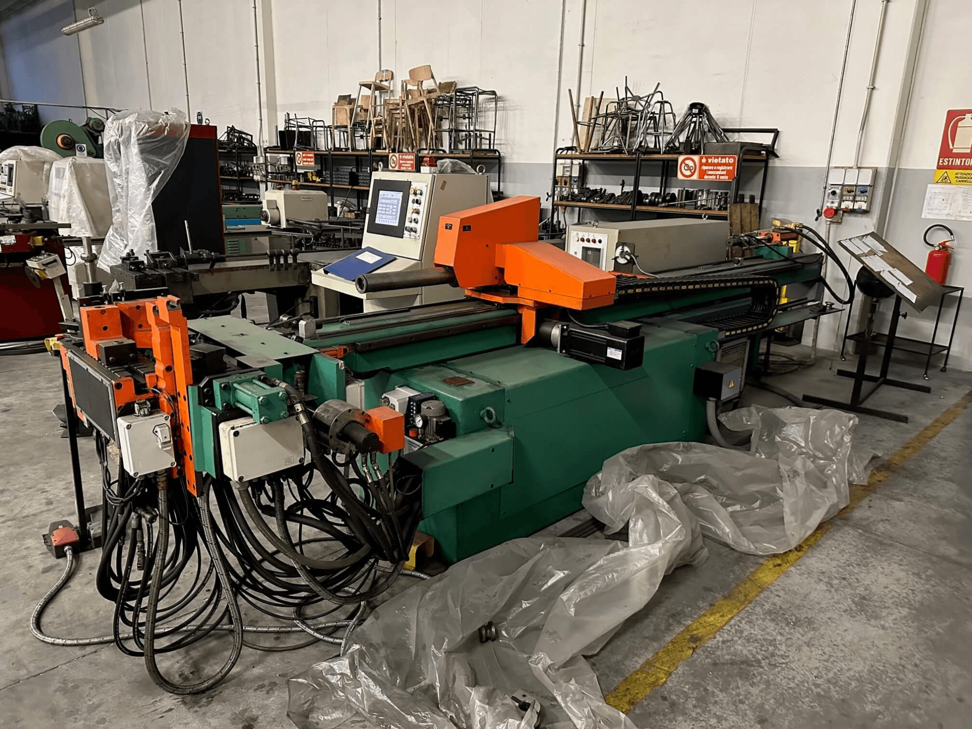 Rūpnieciska mašīna ar oranži zaļu korpusu, vadības paneli un daudziem kabeļiem, skatoties no priekšpuses darbnīcā.