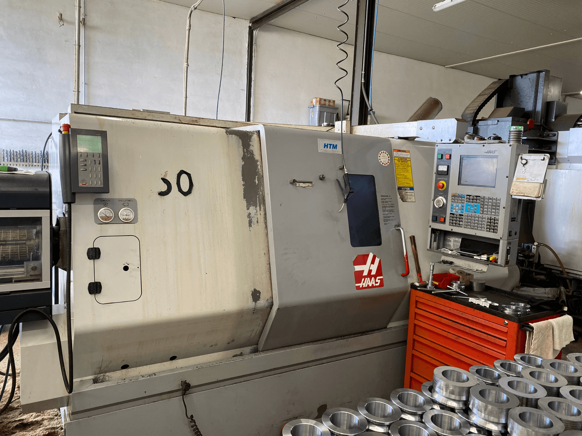 Haas CNC darbgalds ar vadības paneli, skats no priekšpuses, kurā redzamas vadības pogas, mērinstrumenti un blakus sakrautas metāla detaļas.