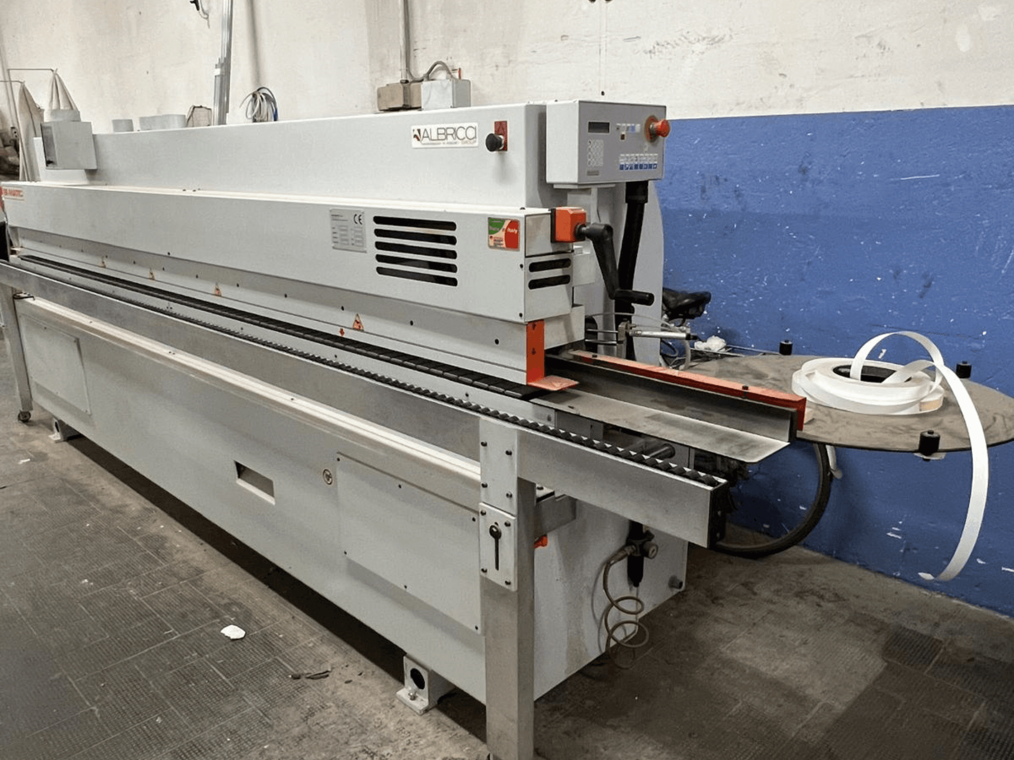 ALBRICO malu aplīmēšanas iekārta no priekšpuses ar garu konveijera lenti un piestiprinātu līmlentes rulli.