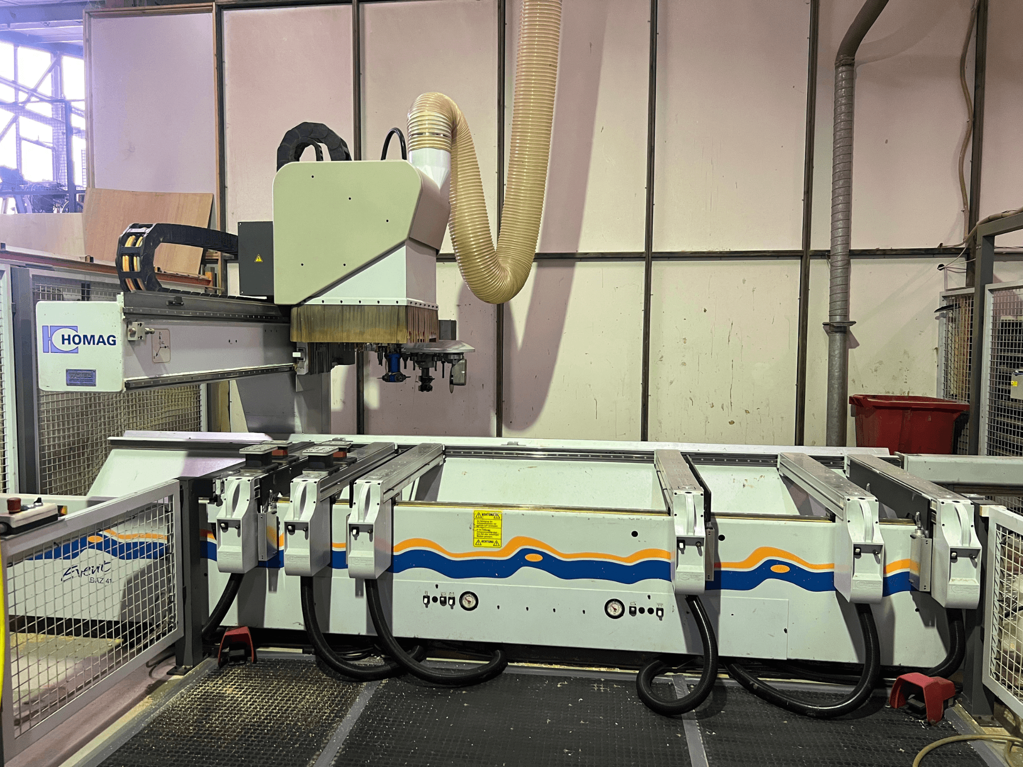 HOMAG BIZ 4 CNC darbnīcā; priekšējais skats, kurā redzama putekļu nosūces sistēma un vadības panelis ar kabeļiem.