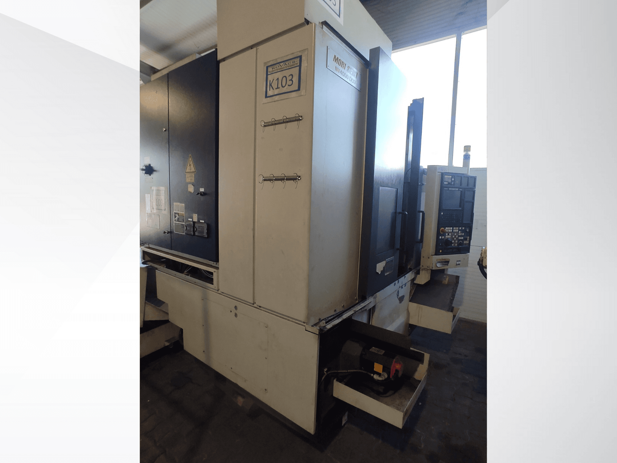 Mori Seiki K103 CNC mašīna priekšpusē, redzams vadības panelis, drošības marķējums un elektriskie komponenti.