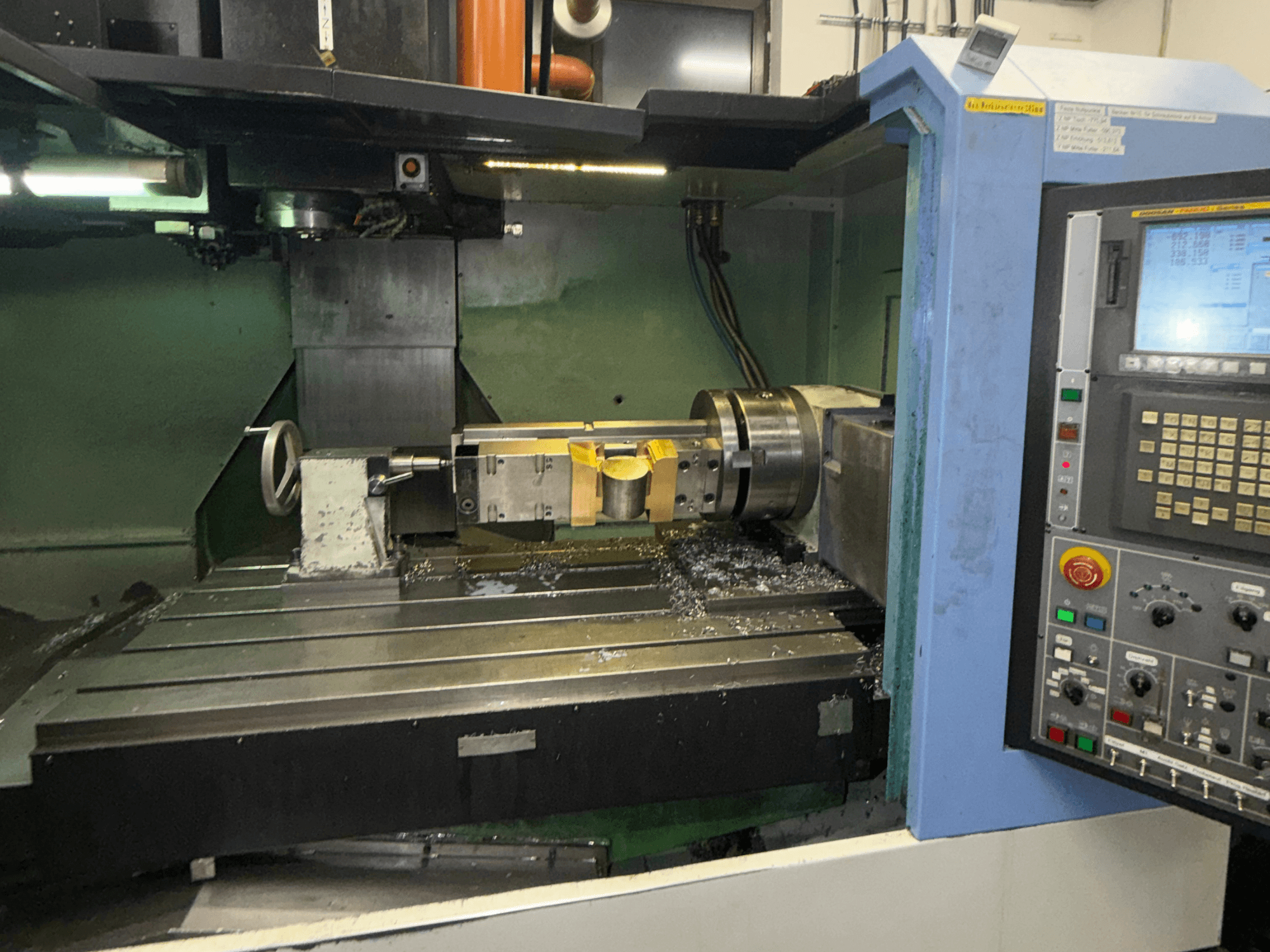 DOOSAN DNM650 vertikālais apstrādes centrs, skats no priekšpuses, kurā redzams griešanas instruments un vadības panelis, kā arī metāla skaidas uz galda.