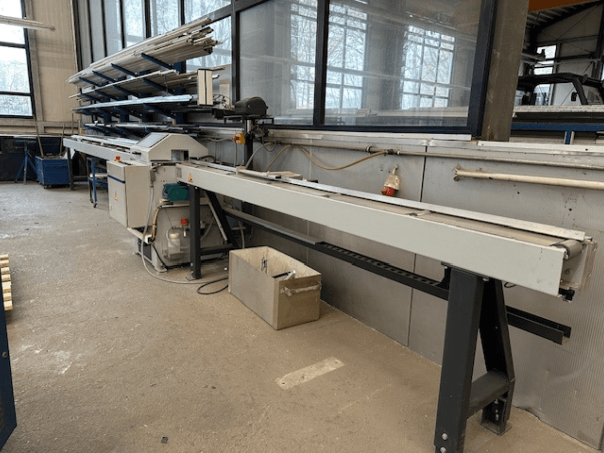 SCHÜCO PGS ripzāģis metālam darbnīcā, skats no sāniem ar regulējamu griešanas gultni un metāla profilu plauktu virs tās.