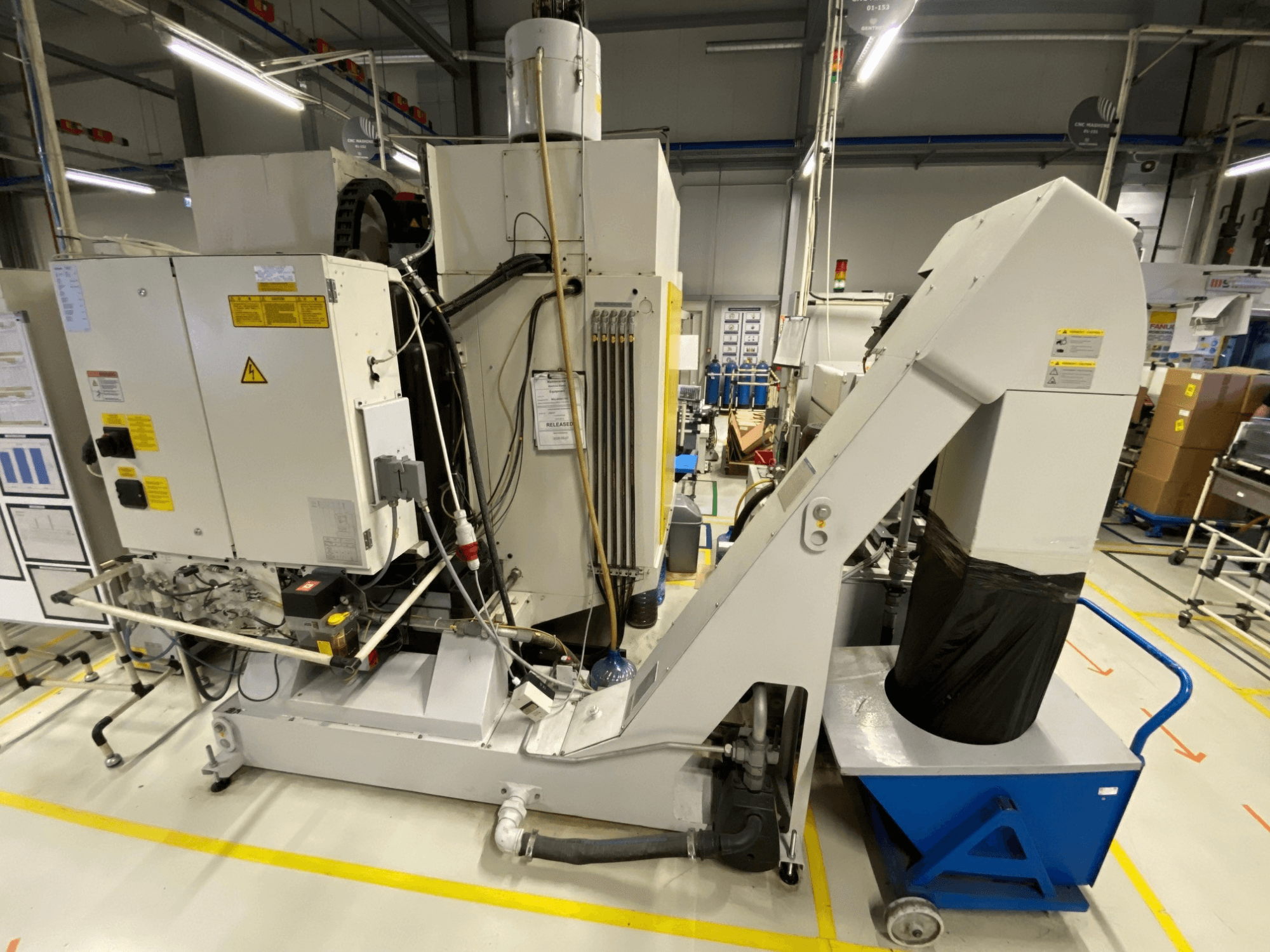 FANUC ROBODRILL α-D21LiB vertikālā apstrādes centra skats no priekšpuses, kurā redzamas elektriskās detaļas un izvadkanāls.