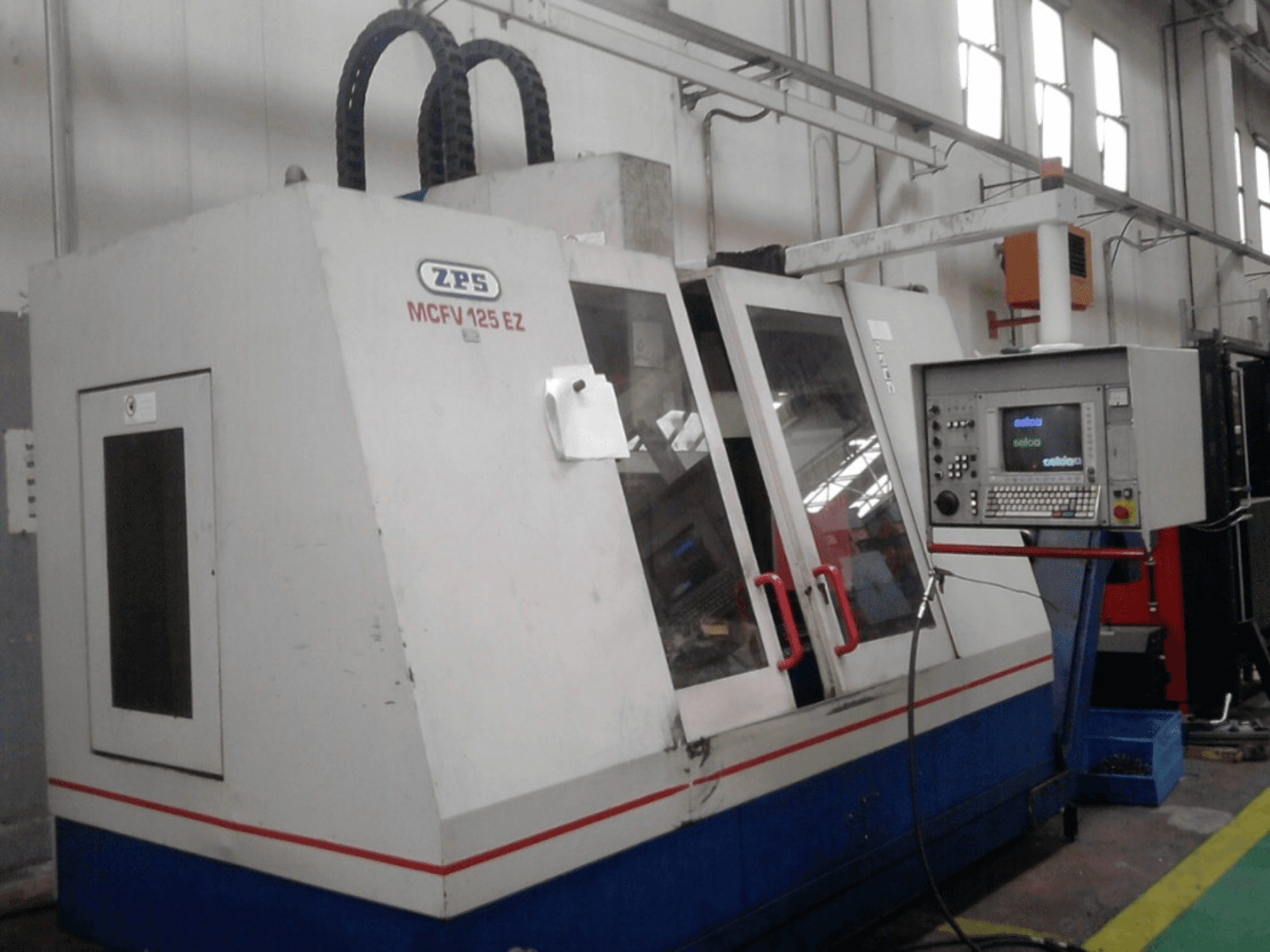 ZPS MCFV 125 EZ CNC mašīna, priekšējais skats ar vadības paneli un sarkaniem rokturiem, darbnīcā ar redzamām šļūtenēm.
