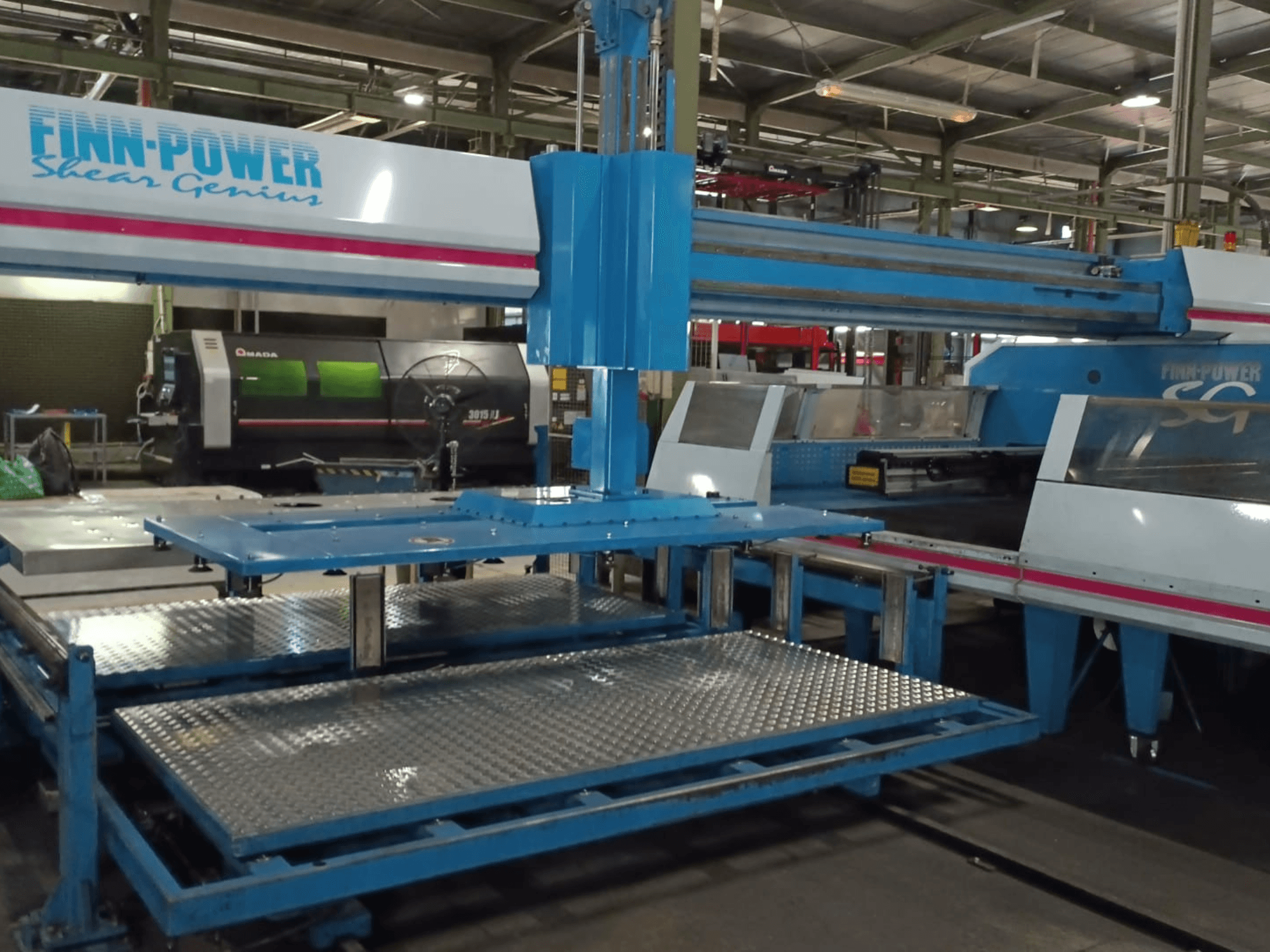 Finn-Power SG6 „Shear Genius“ CNC perforācijas mašīna, skats no priekšpuses, kurā redzami metāla galdi un konstrukcijas elementi.