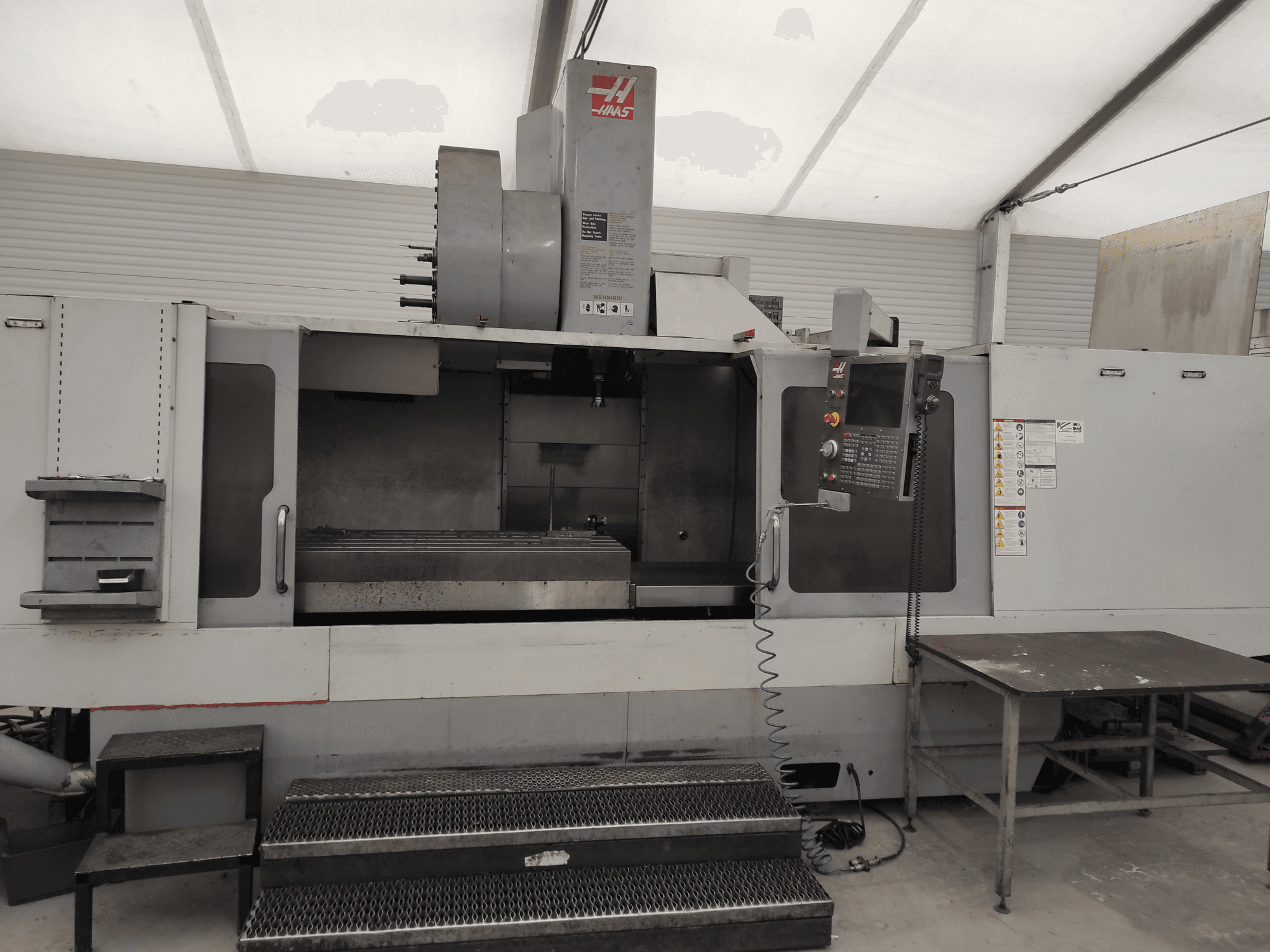 HAAS VF-10/50HE vertikālās apstrādes centrs, priekšējais skats, kurā redzams vadības panelis, darbgalda un darbam pieejamais pakāpiens.