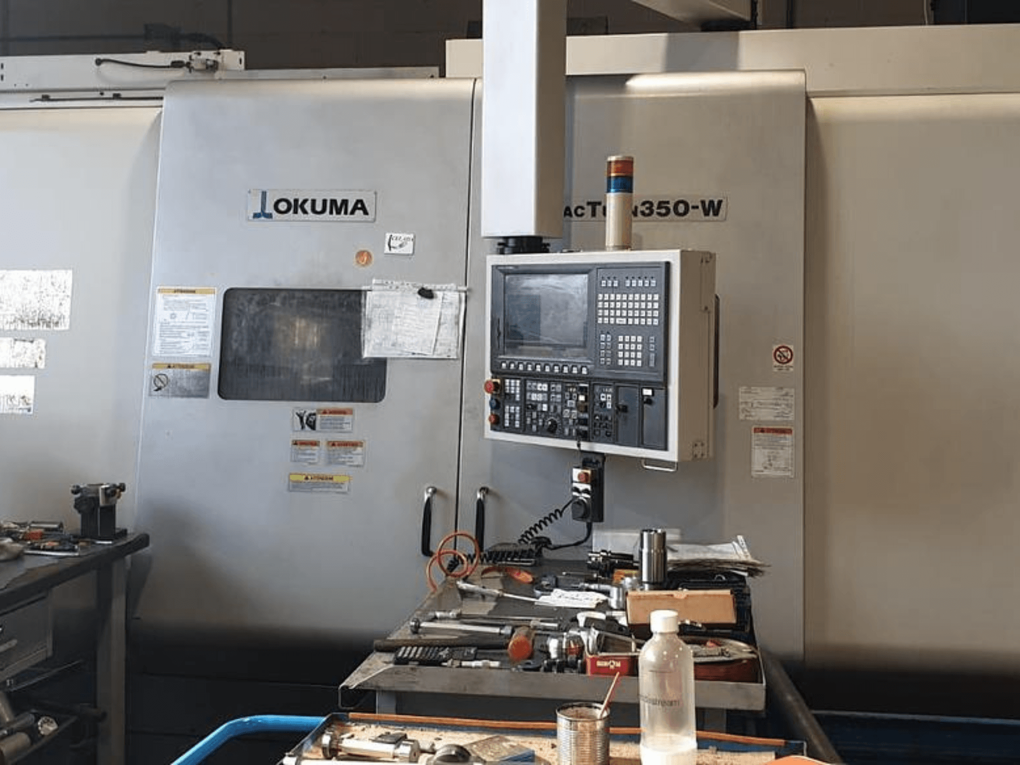 Okuma MAC TURN 350-W frēzēšanas centrs, skats no priekšpuses ar vadības paneli un darba vietu, kas pārpildīta ar instrumentiem un pudeli.