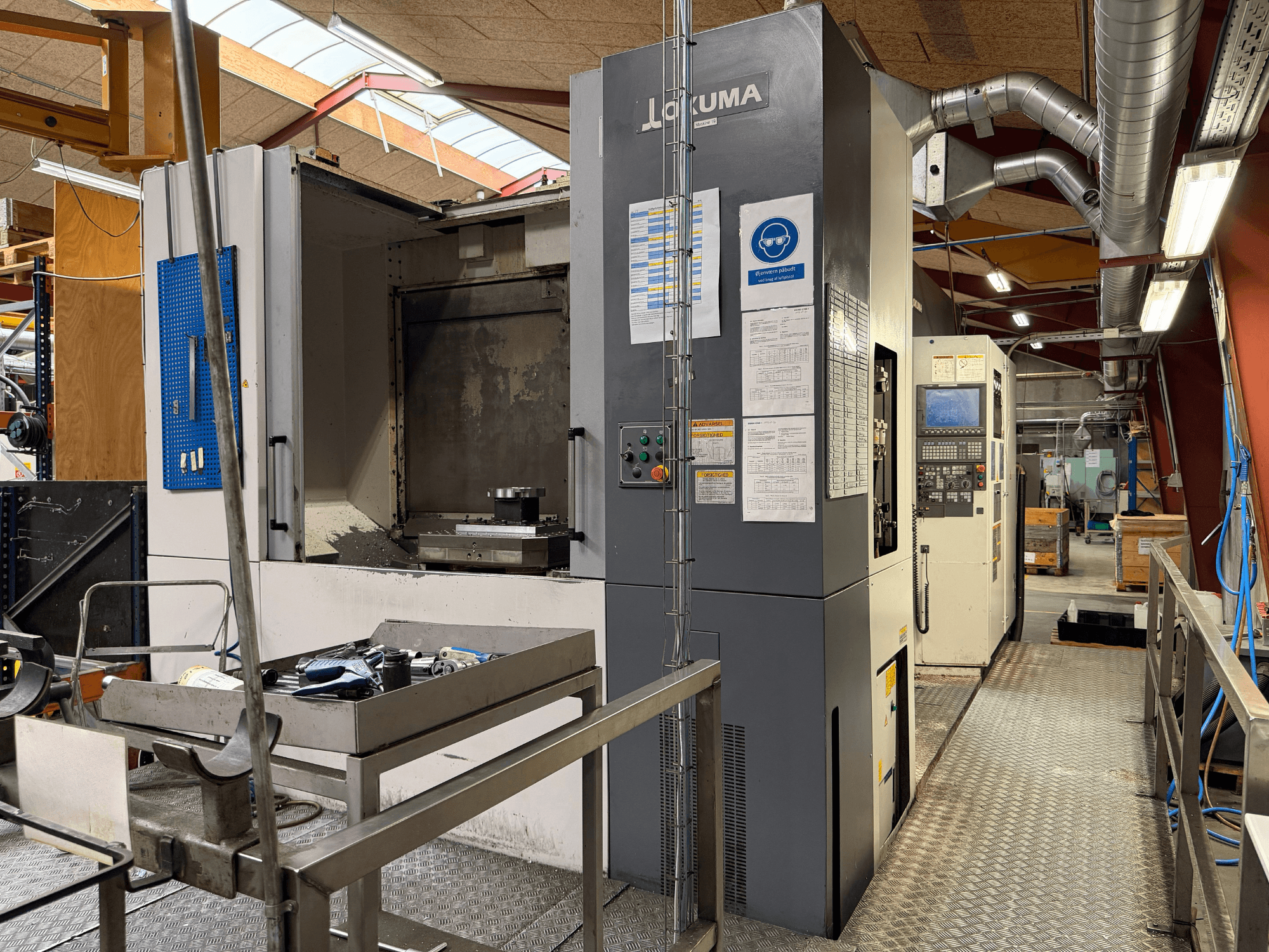 Okuma MA-600HB horizontālais apstrādes centrs, skats no priekšpuses, kurā redzami instrumenti uz metāla galda un mašīnas vadības elementi.