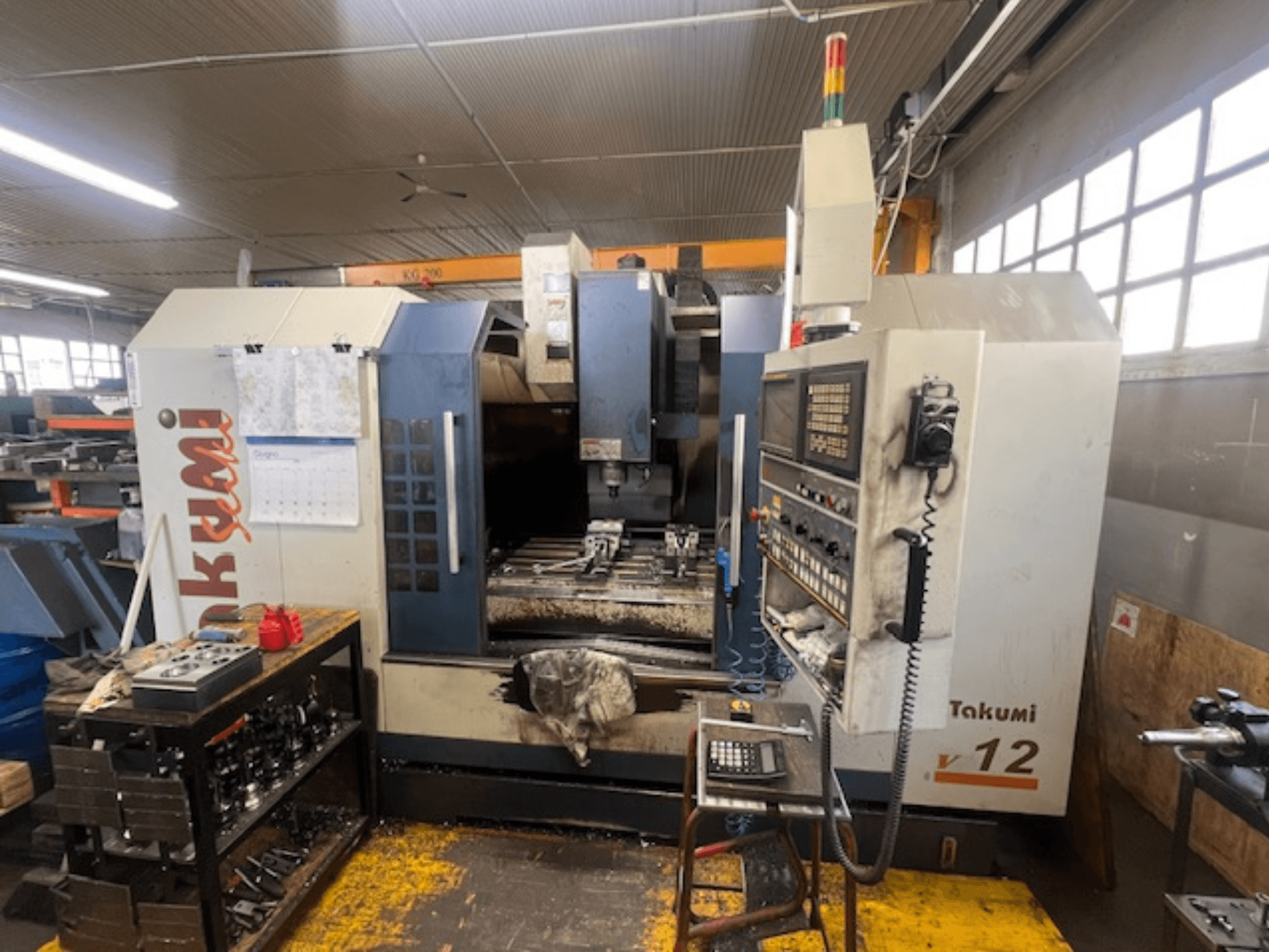 Takumi V12 CNC frēzmašīnas sānu skats, kurā redzams vadības panelis, griešanas zona un darbstacijas instrumenti.