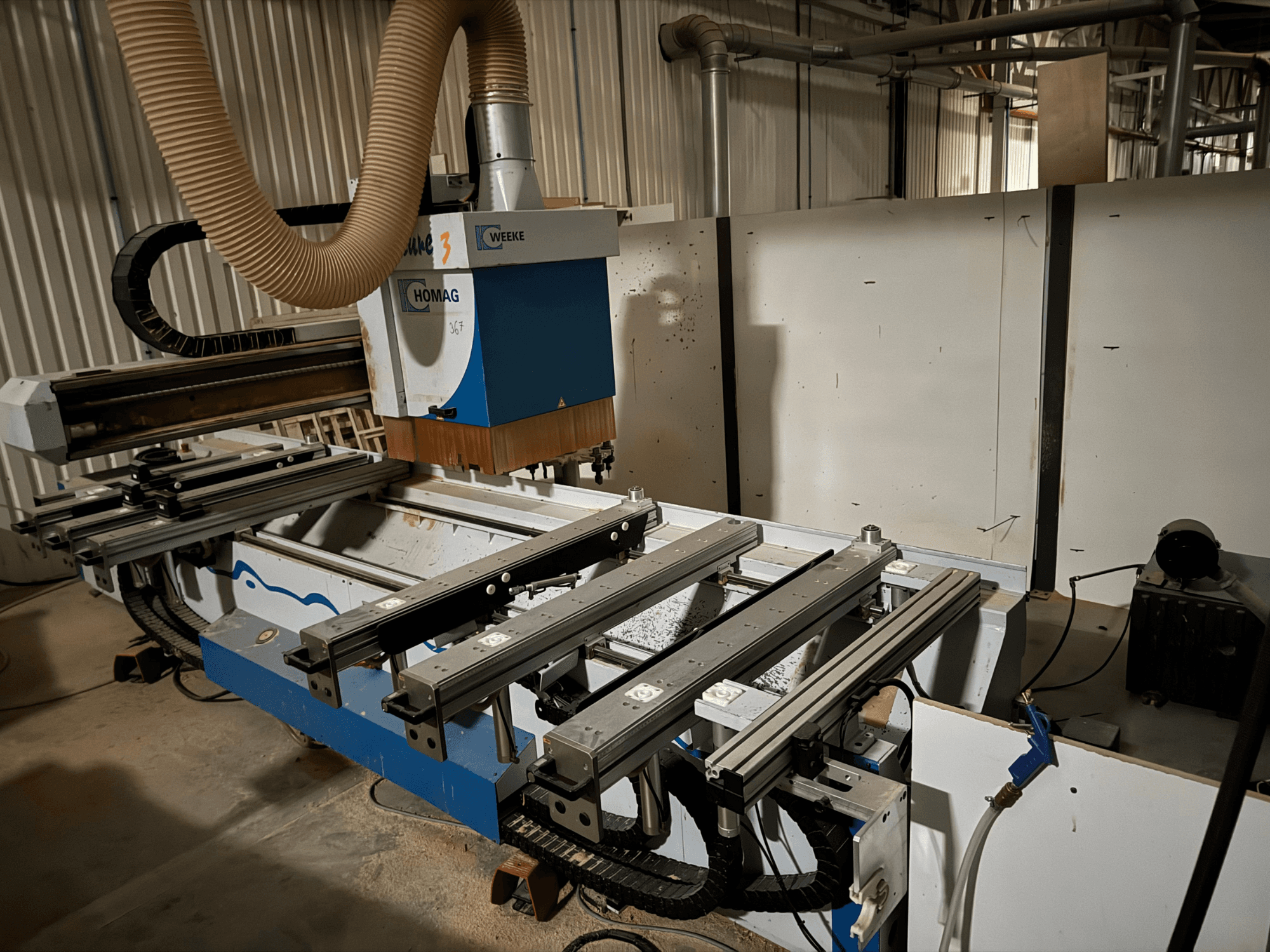 HOMAG CNC darbgalds zilā un baltā krāsā, skats no sāniem, aprīkots ar bīdāmām sliedēm, putekļu nosūces sistēmu un piestiprinātiem instrumentiem.