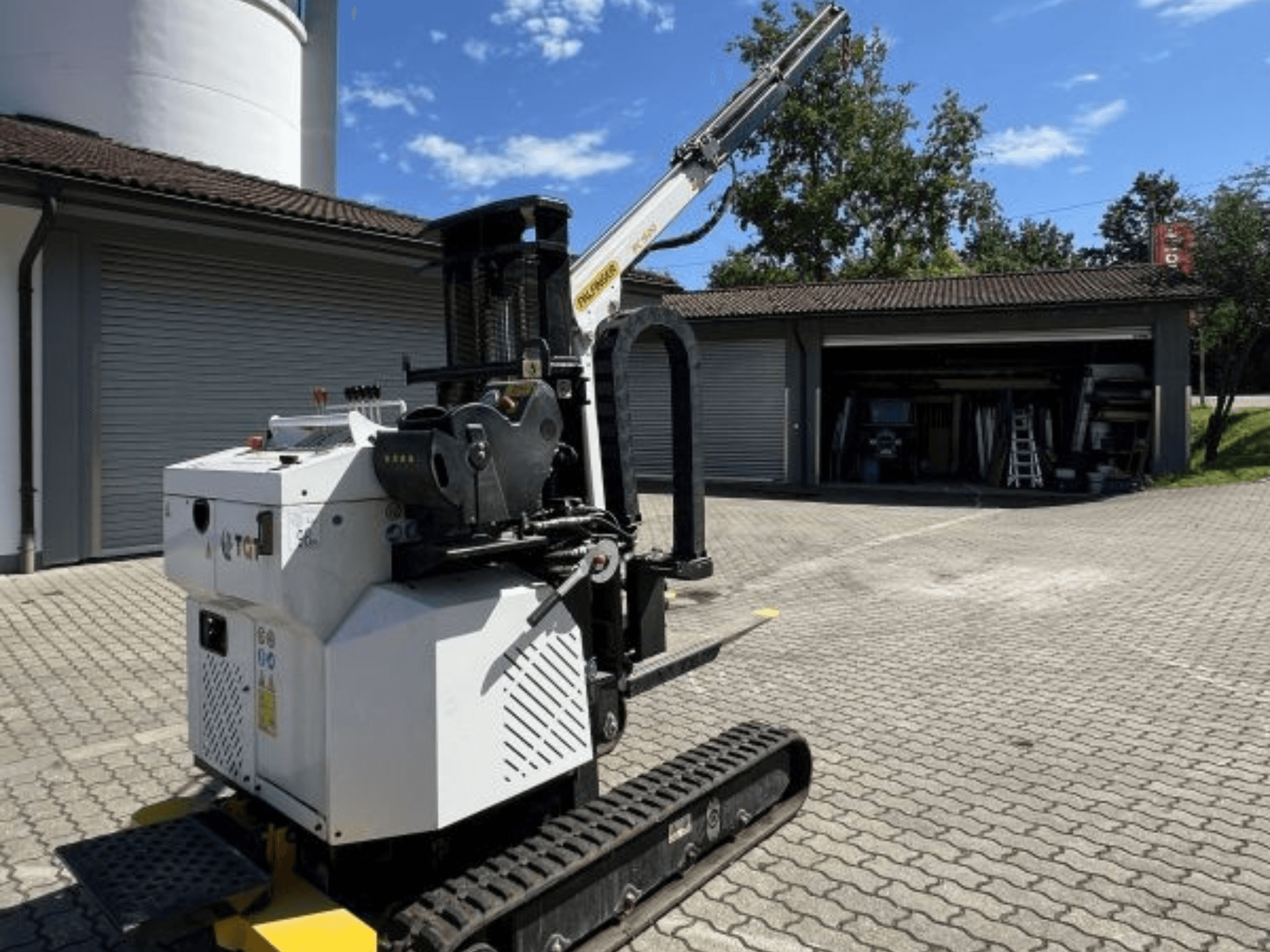 TGI celtniecības mini celtnis, skats no sāniem, redzamas kāpurķēdes un uzmontēta strēle, novietots uz ķieģeļu virsmas pie garāžas.