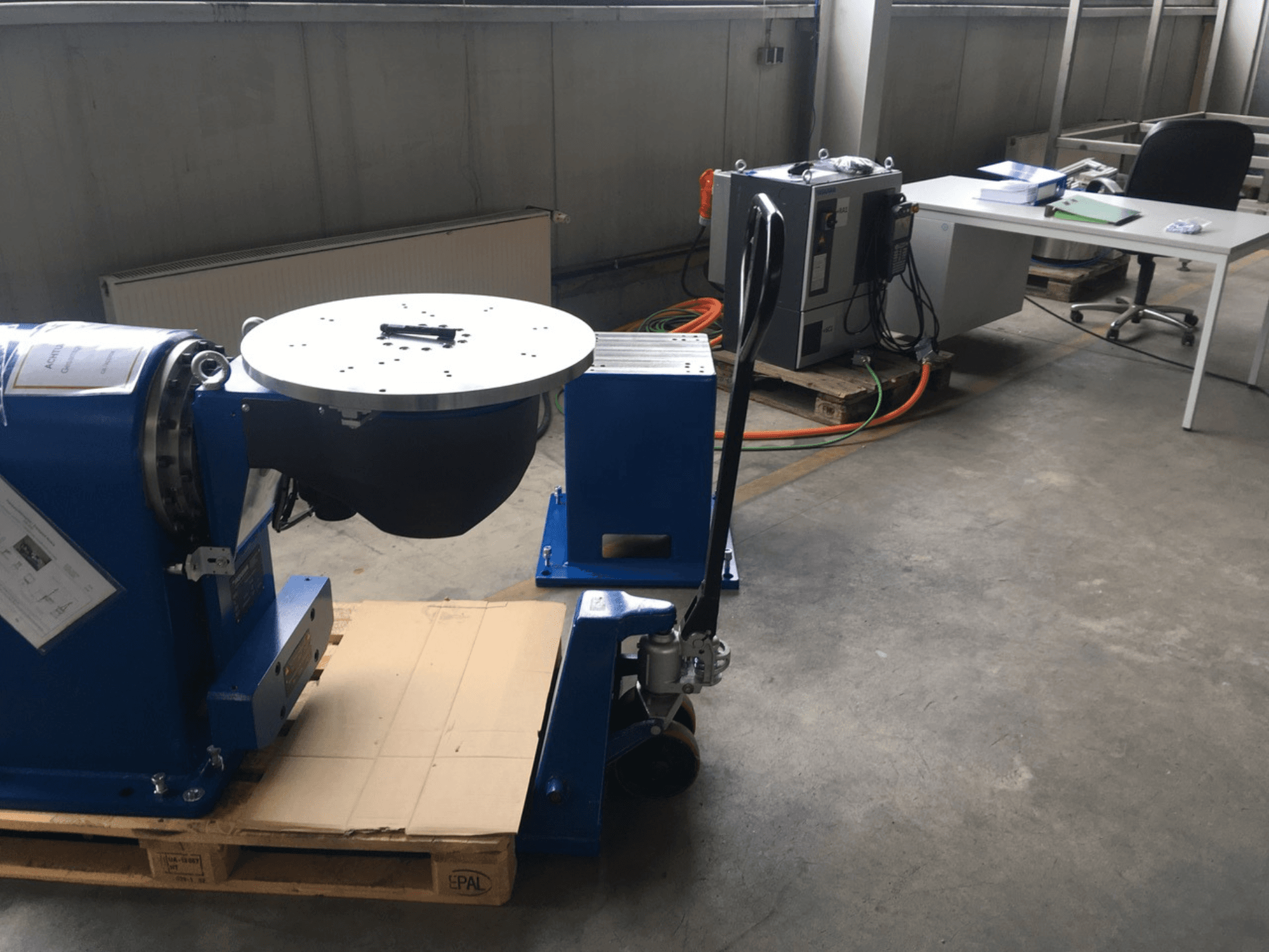 Zila rūpnieciska iekārta ar apaļu pamatni un vadības paneli, skatoties no priekšpuses, uz koka paliktņa birojā.