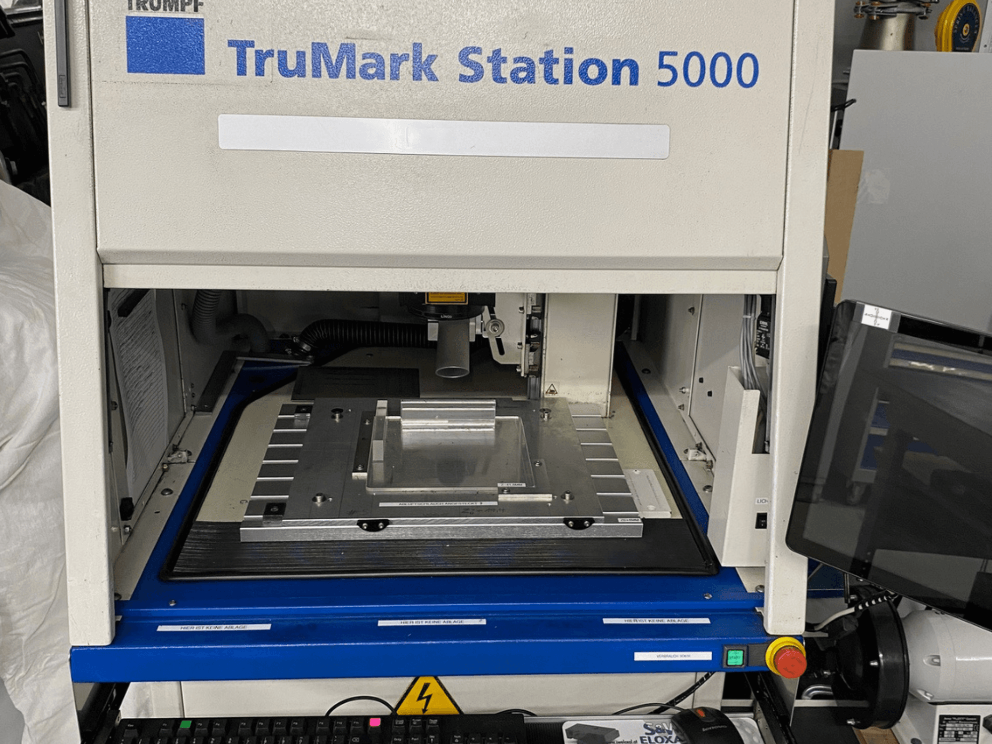 TruMark Station 5000 lāzera marķēšanas iekārtas priekšējais skats, kurā redzams darba galds, vadības ierīces un uzraudzības ekrāns.