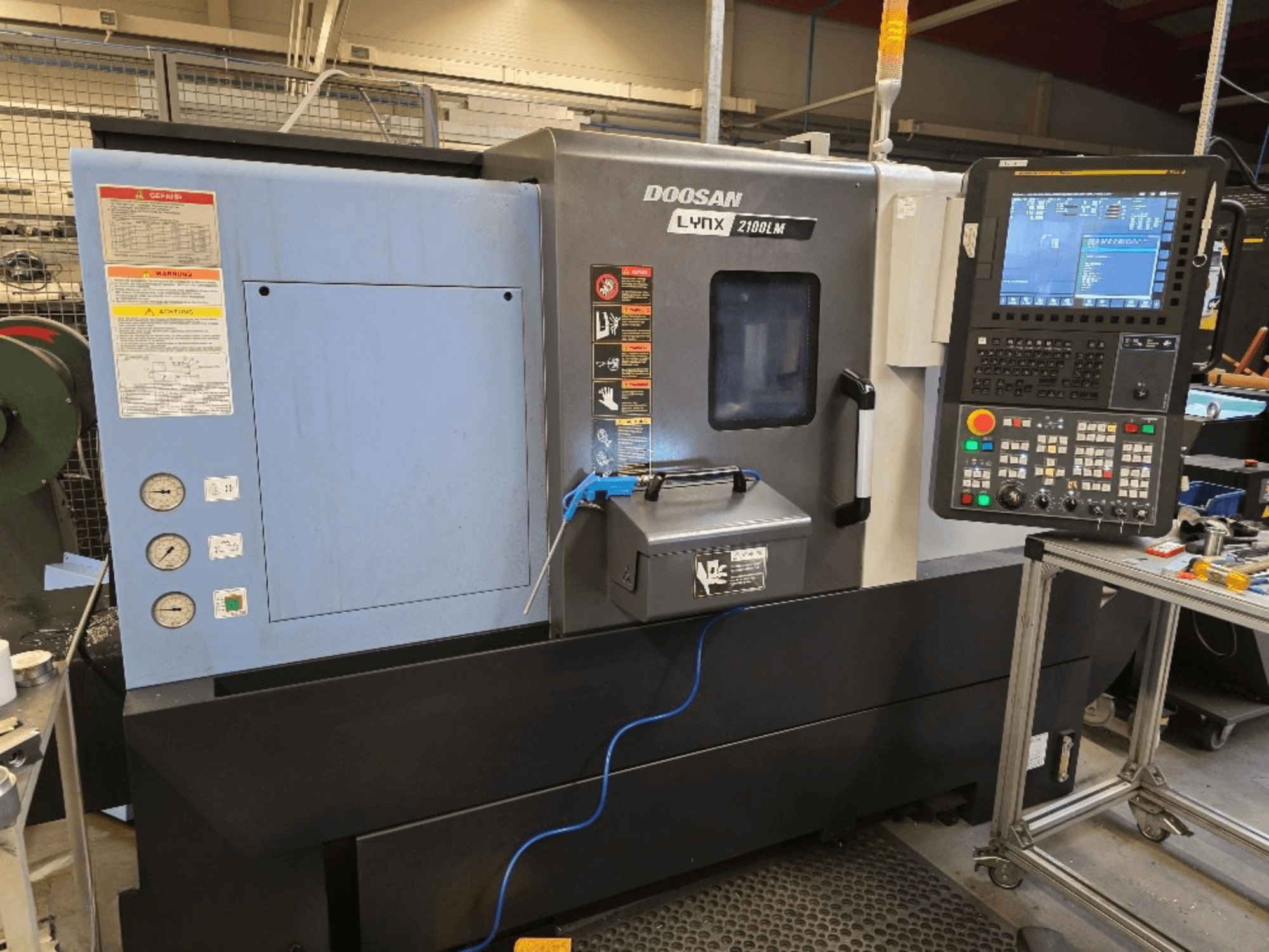 Doosan Lynx 2100LM CNC virpa, skats no priekšpuses, ar vadības paneli, spiediena mērinstrumentiem un drošības brīdinājumiem.