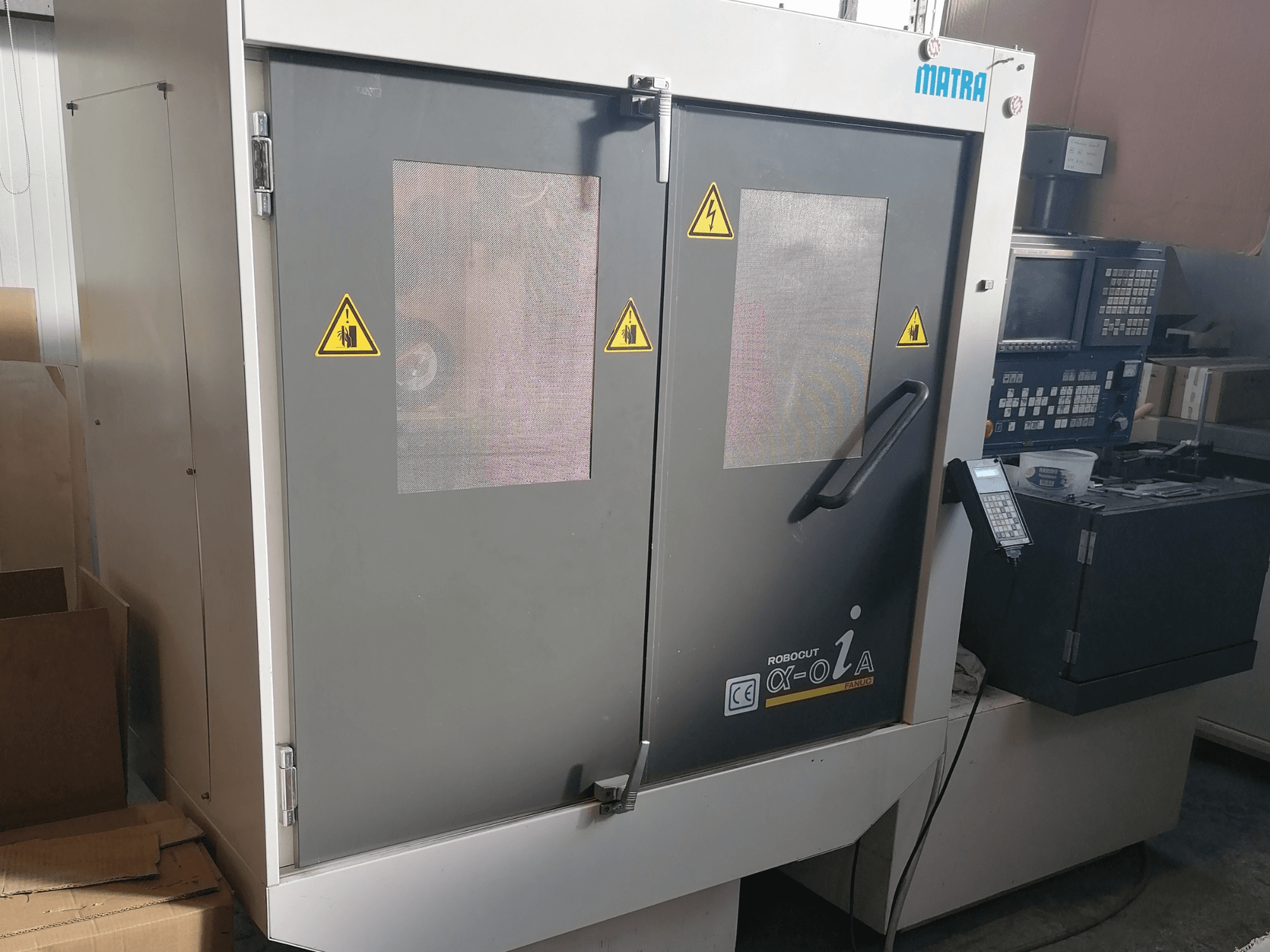 Matra Robocut α-oZα mašīna, priekšējais skats, kurā redzami drošības brīdinājumi, vadības panelis un dubultās durvis ar režģa logiem.