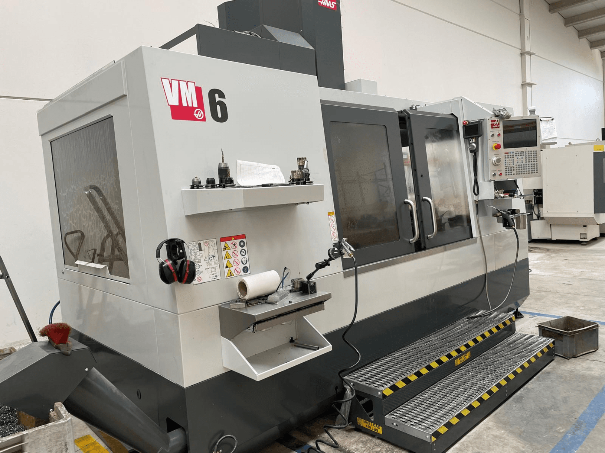 Haas VM-6 vertikālais apstrādes centrs, priekšējais skats, kurā redzams vadības panelis, instrumentu turētāji un drošības elementi.