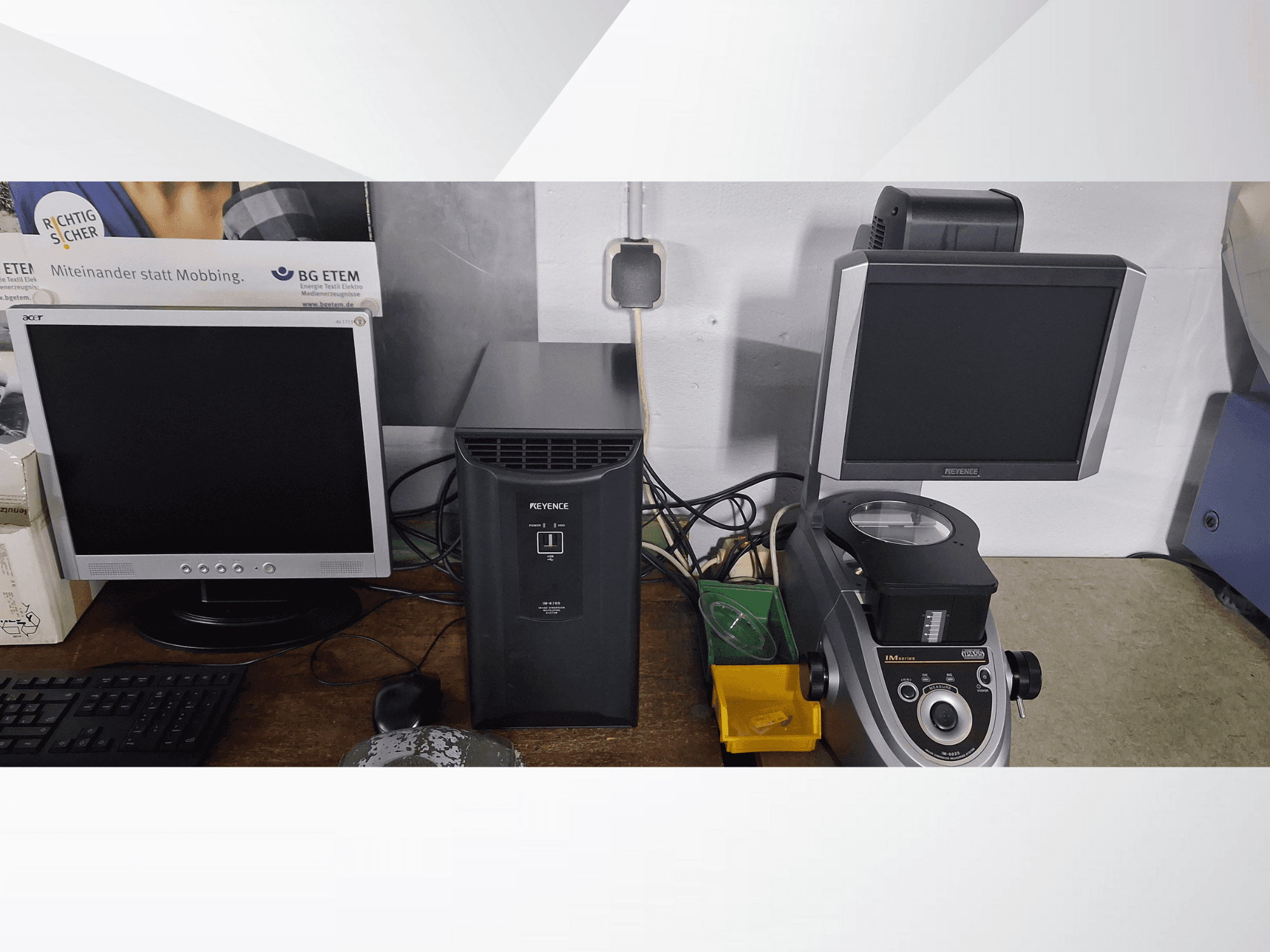 Divi datora monitori, viens Acer un viens Keyence, blakus melnai ierīcei un mikroskopam, skatoties no priekšpuses.