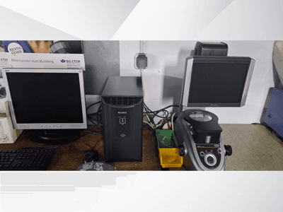 Divi datora monitori, viens Acer un viens Keyence, blakus melnai ierīcei un mikroskopam, skatoties no priekšpuses.