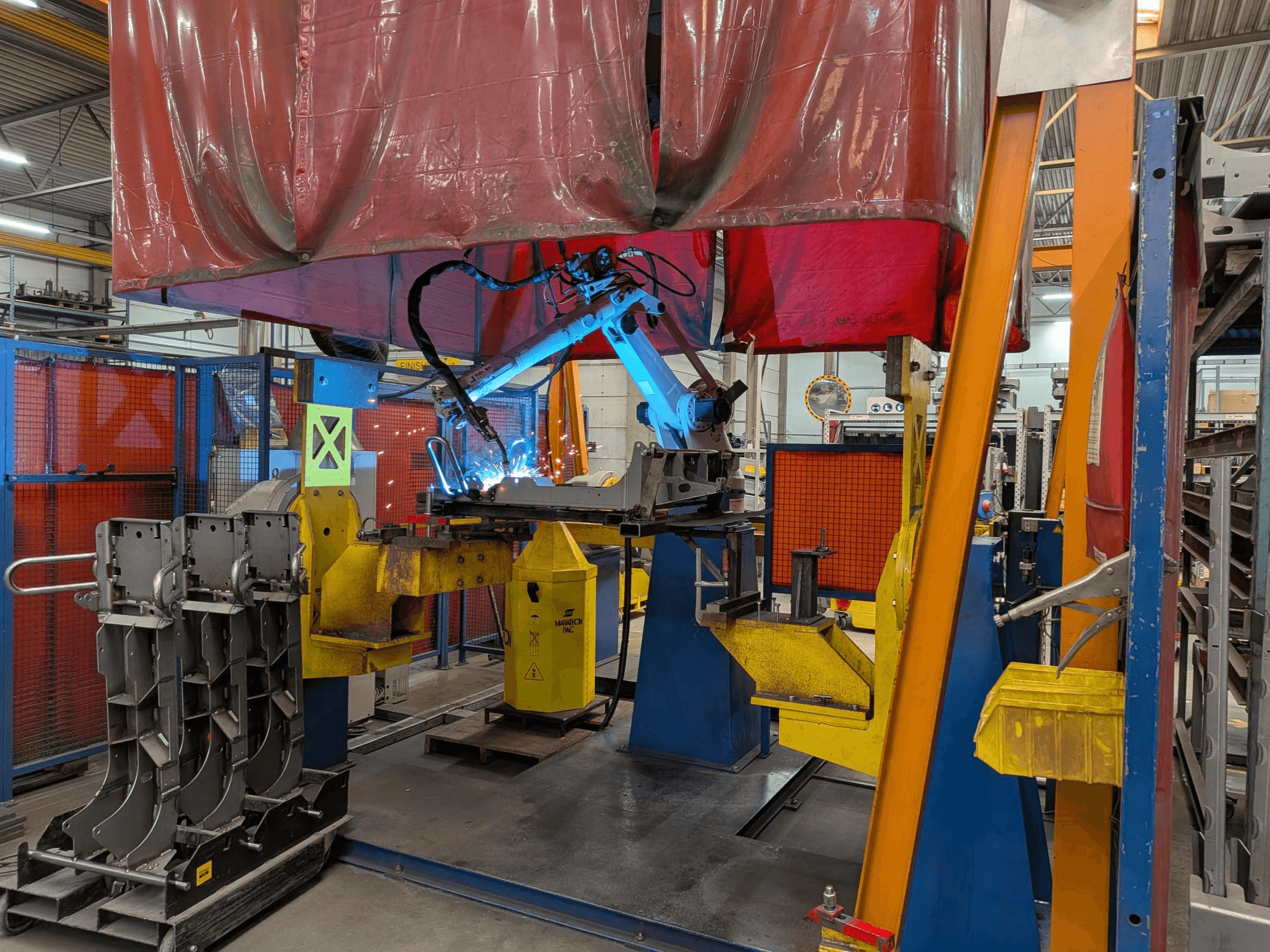 Metināšanas robots OTC Daihen DR4200 darbībā, skats no priekšpuses, ar spilgti zilu metināšanas loku un aizsargpārsegu.
