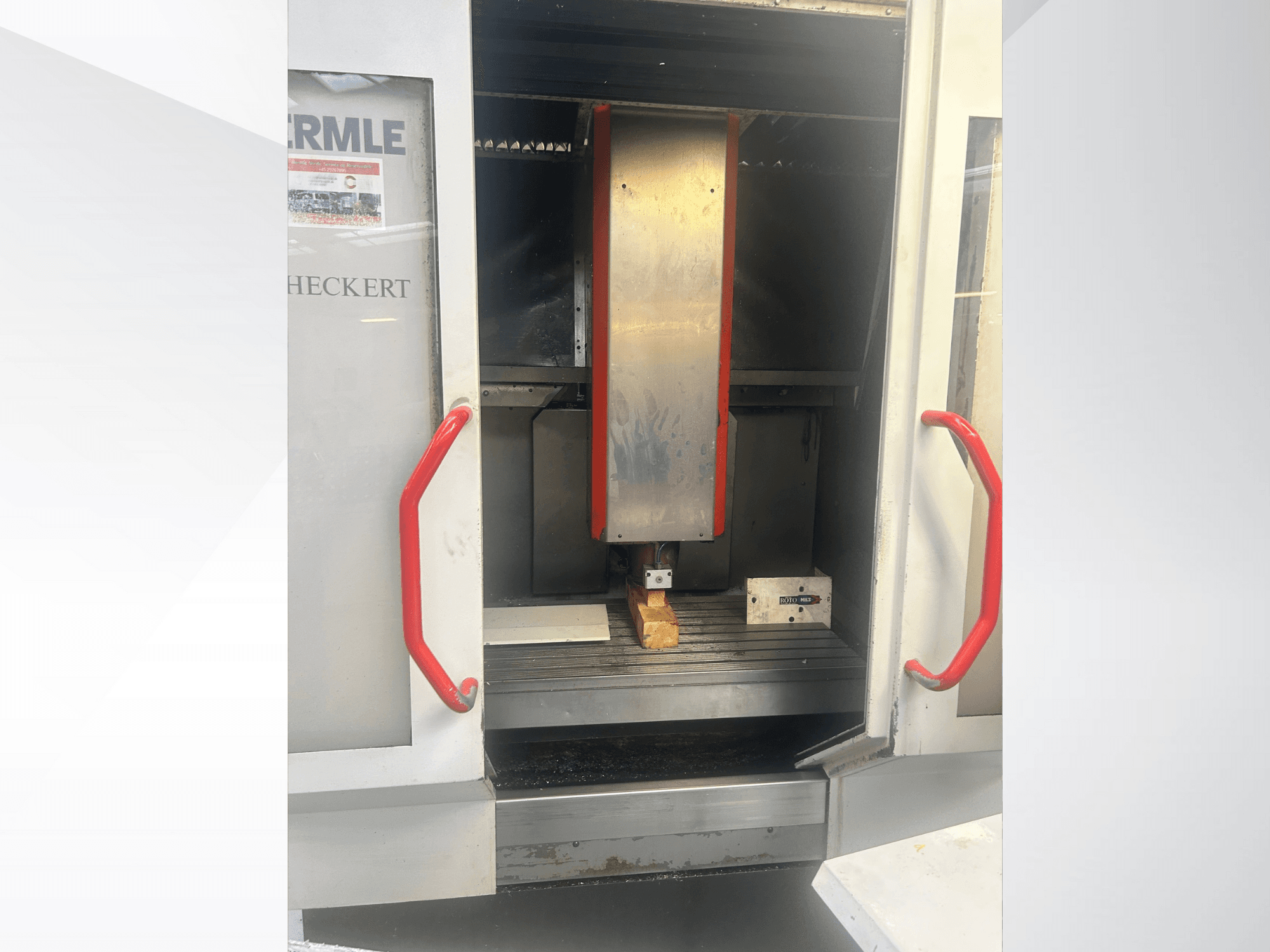 Hermle CNC mašīnas priekšējais skats ar metālisku iekšpusi, sarkaniem rokturiem un apstrādājamo detaļu uz galda.