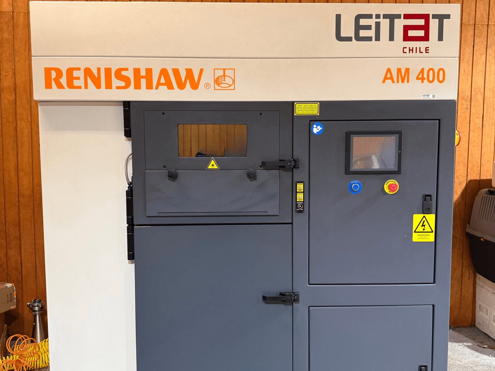 RENISHAW AM400 metāla 3D printera skats no priekšpuses, kurā redzams vadības panelis, brīdinājuma uzlīmes un piekļuve apkopes veikšanai.
