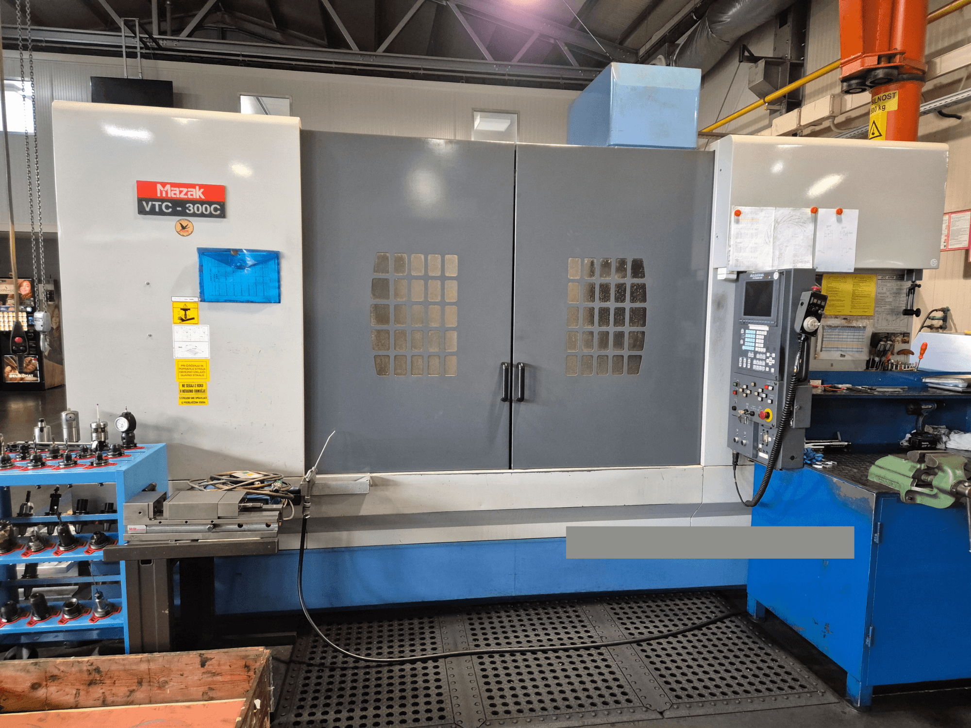 Mazak VTC-300C mašīna rūpnīcā, priekšējais skats, kurā redzams vadības panelis, pelēkas durvis ar logu režģi un tuvumā esošās iekārtas.