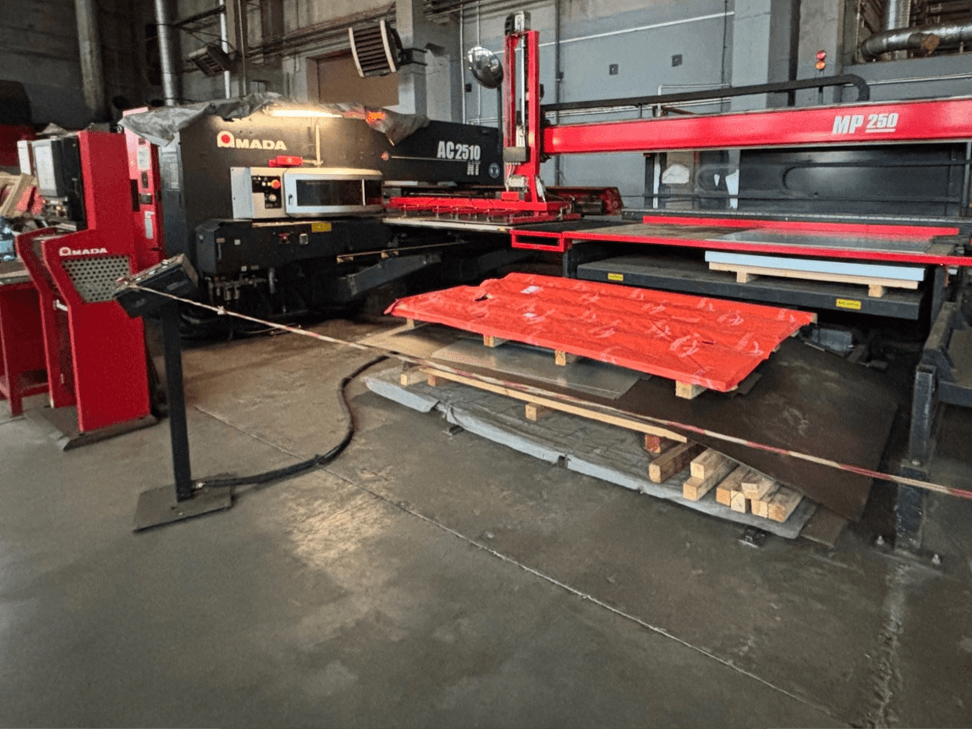 Amada AC2510 NT lāzera griezējs rūpnīcā, skats no priekšpuses, smagās iekārtas ar metāla loksnēm un paletēm uz grīdas.