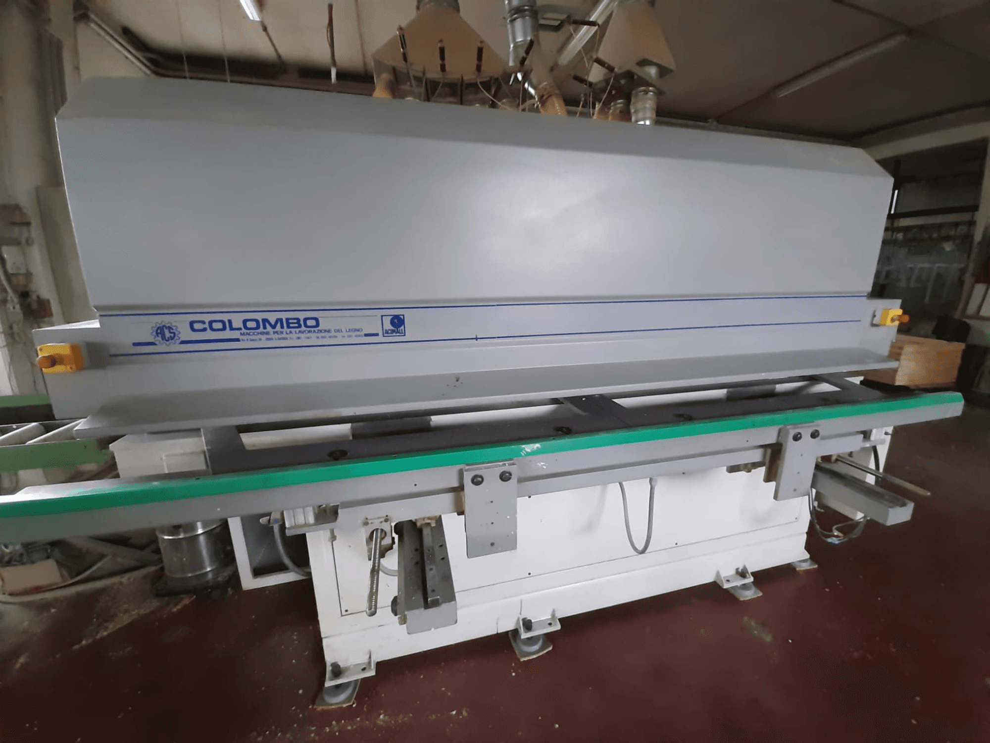Colombo kokapstrādes mašīna, skats no priekšpuses, ar garu zaļu galdu, vadības pogām un pelēku korpusu ar ziliem akcentiem.