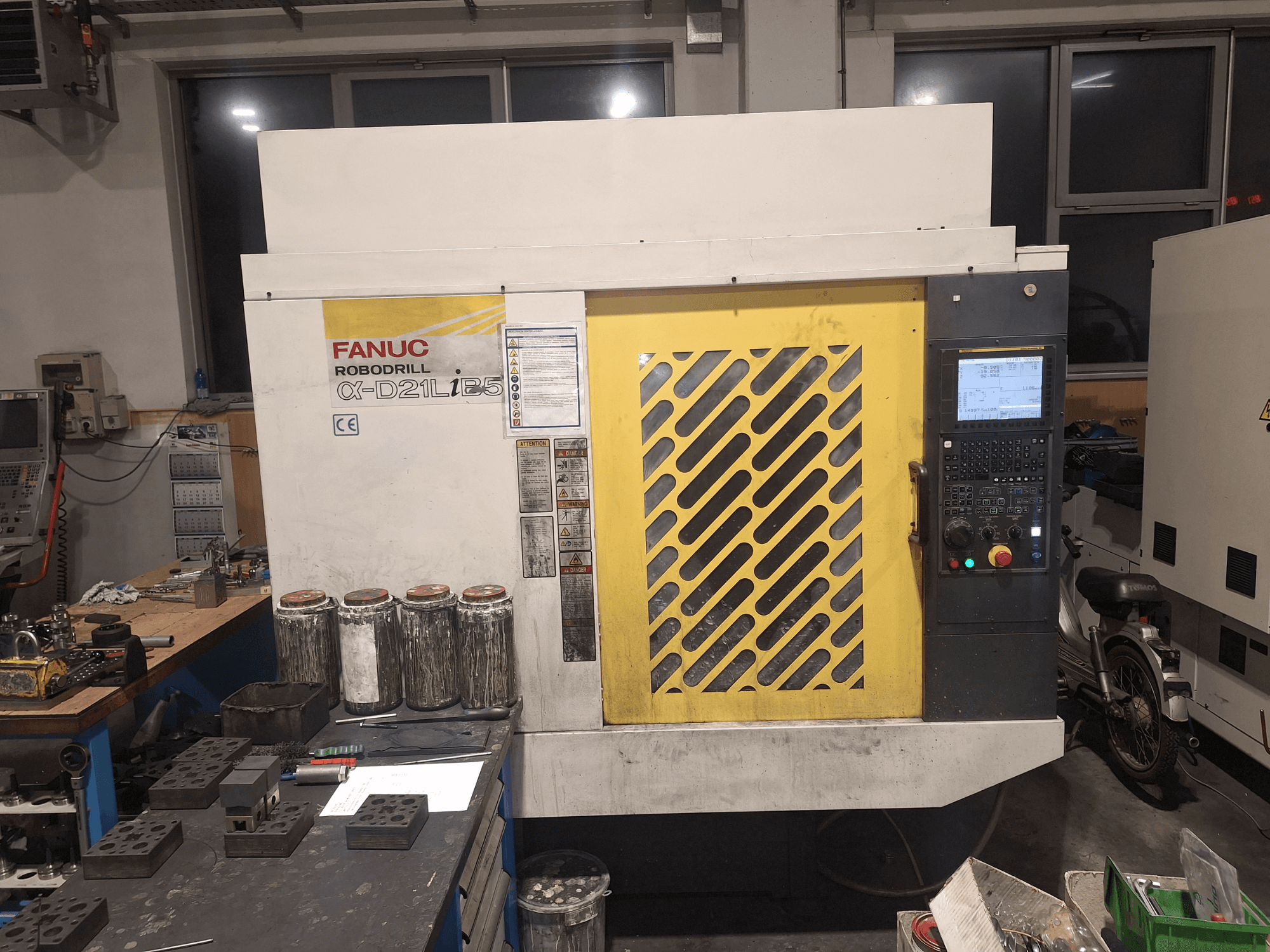 FANUC Robodrill α-D21LiB5 priekšējais skats, kurā redzams vadības panelis, dzeltenā režģa un tuvumā esošie darbgaldā novietoti instrumenti.