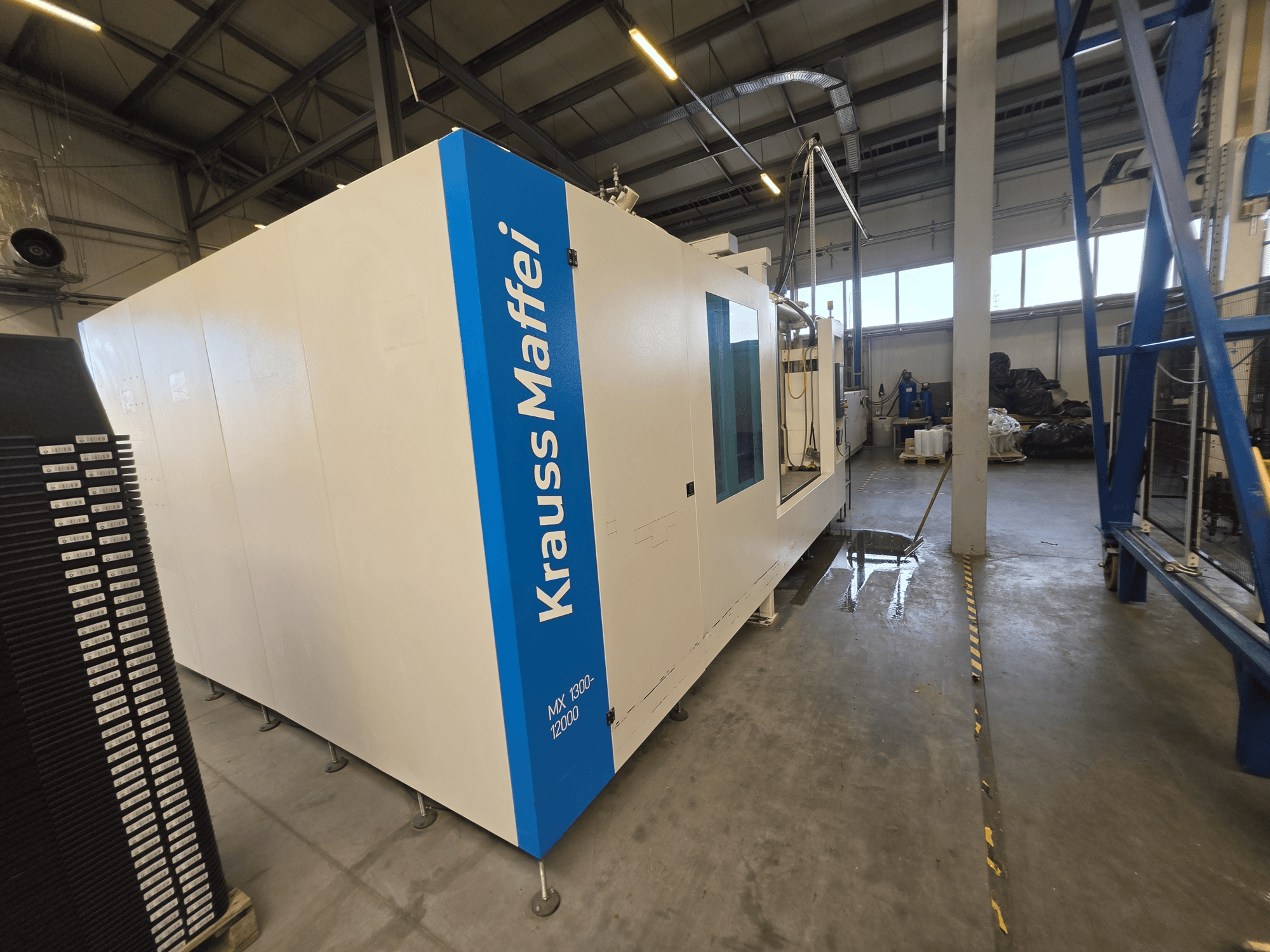 Krauss Maffei MX 1300-12000 hidrauliskā injekcijas formēšanas mašīna no sāniem, kurā redzams tās lielais baltais šasijas korpuss un zils zīmola logotips.