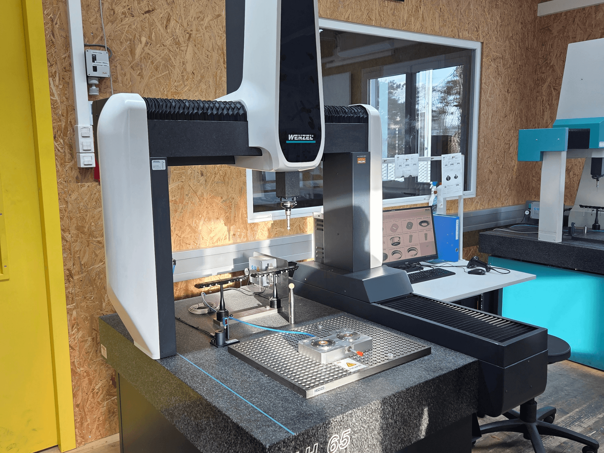 Wenzel LH 65 koordinātu mērīšanas iekārta laboratorijas uzstādījumā, skats no priekšpuses, uz kuras redzami mērinstrumenti un monitors.