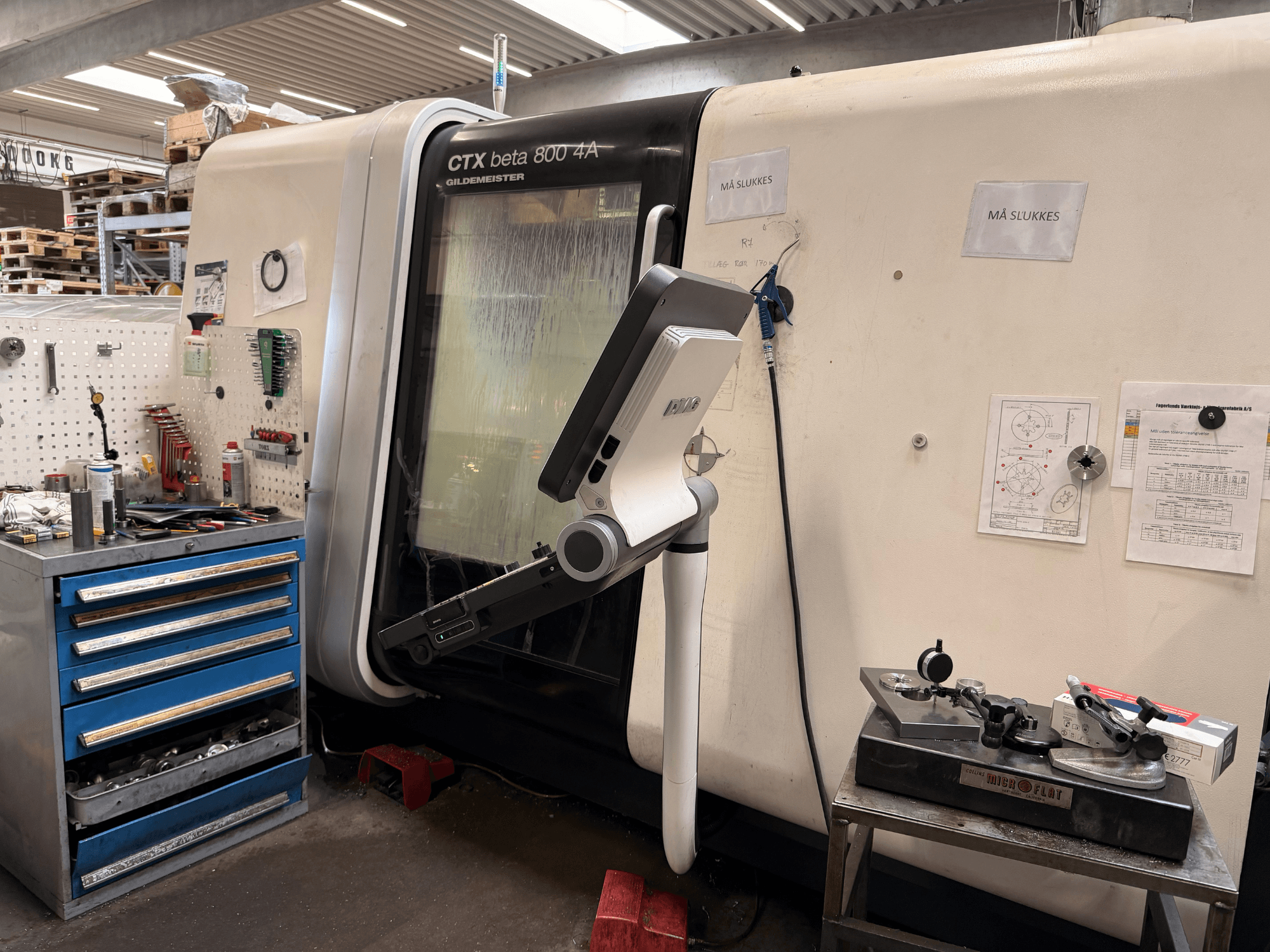 DMG MORI CTX Beta 800 4a vertikālās virpošanas mašīna darbnīcā, ar vadības roku un tehnisko dokumentāciju blakus.