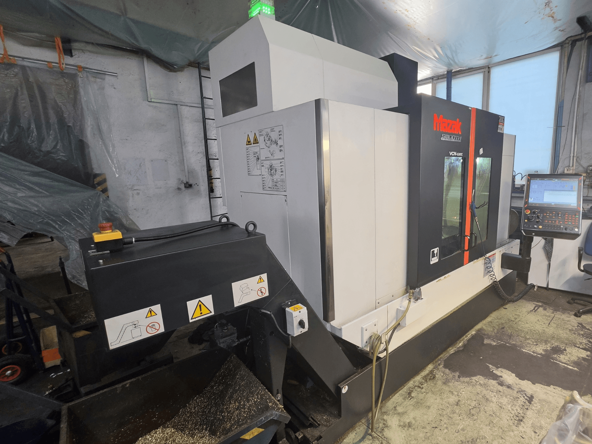 Mazak VCN-530C vertikālā apstrādes centra skats no priekšpuses, kurā redzams vadības panelis, instrumentu ķēde un darbības indikatori.