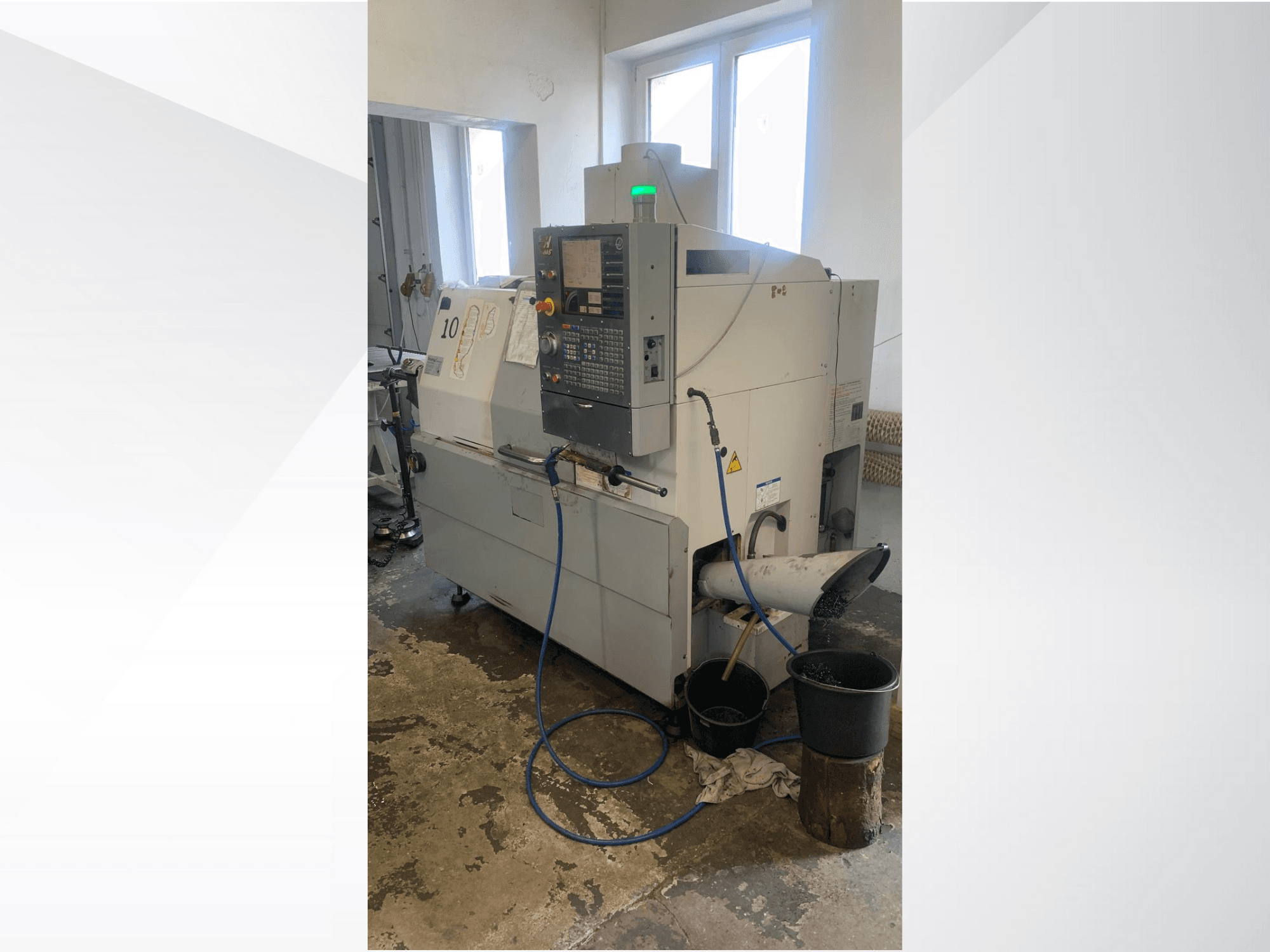 Haas CNC darbgaldu modelis ar vadības paneli, skats no sāniem, ar dzesēšanas šķidruma padeves līniju, atkritumu izlaidi un uzglabāšanas tvertnēm apakšā.
