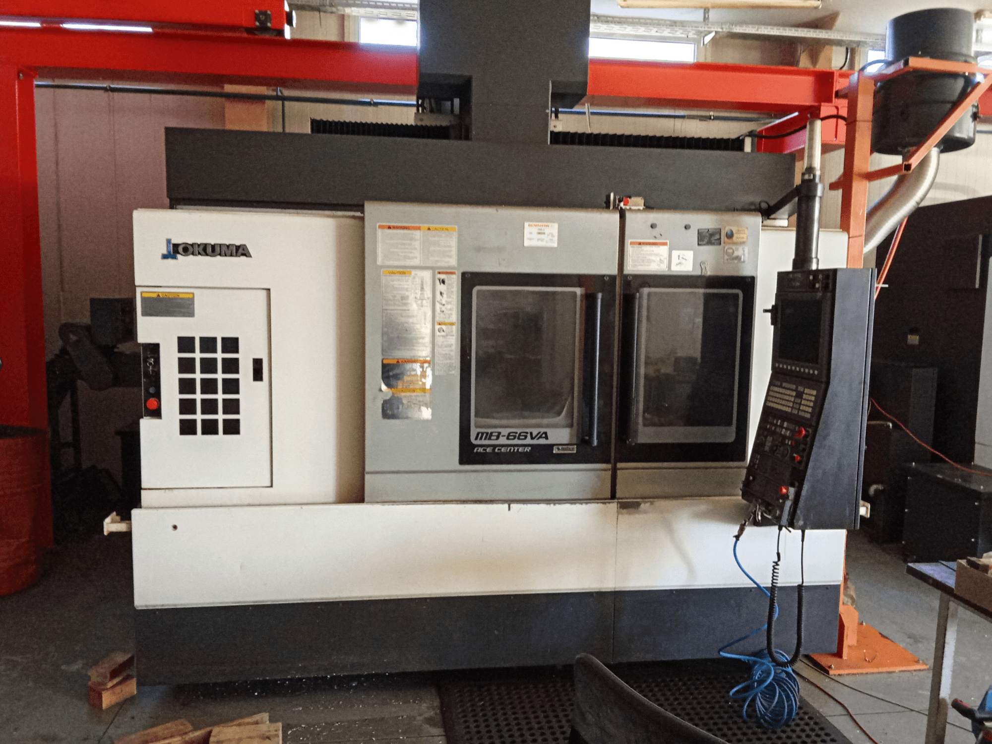 Okuma MB-66VA vertikālais apstrādes centrs, priekšējais skats, kurā redzams vadības panelis un divi skatu logi instrumentu piekļuvei.