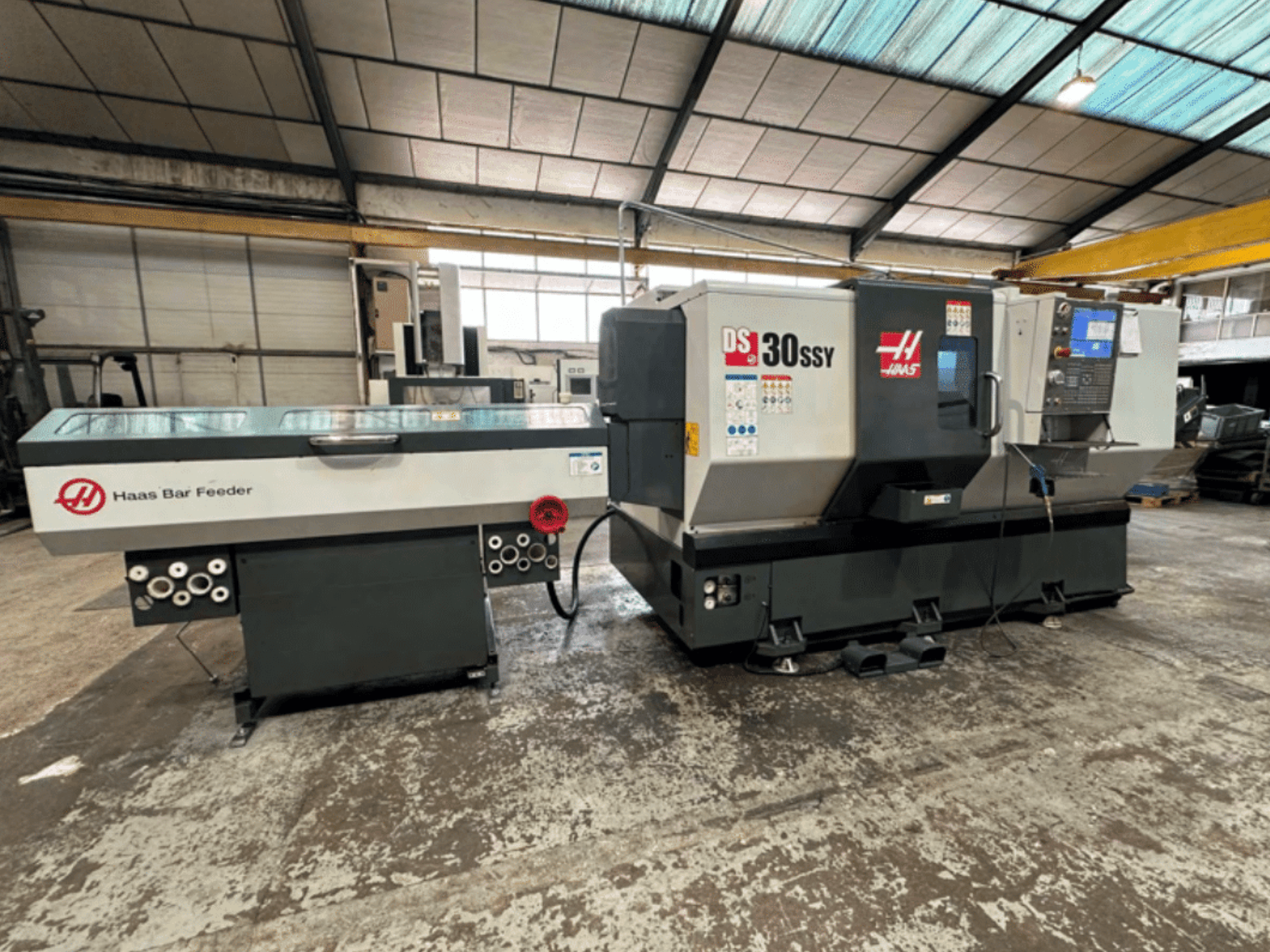 Haas DS-30SSY CNC virpa ar Haas stieņu padevēju, sānu skats, kurā redzams vadības panelis un padeves mehānisms darbnīcā.