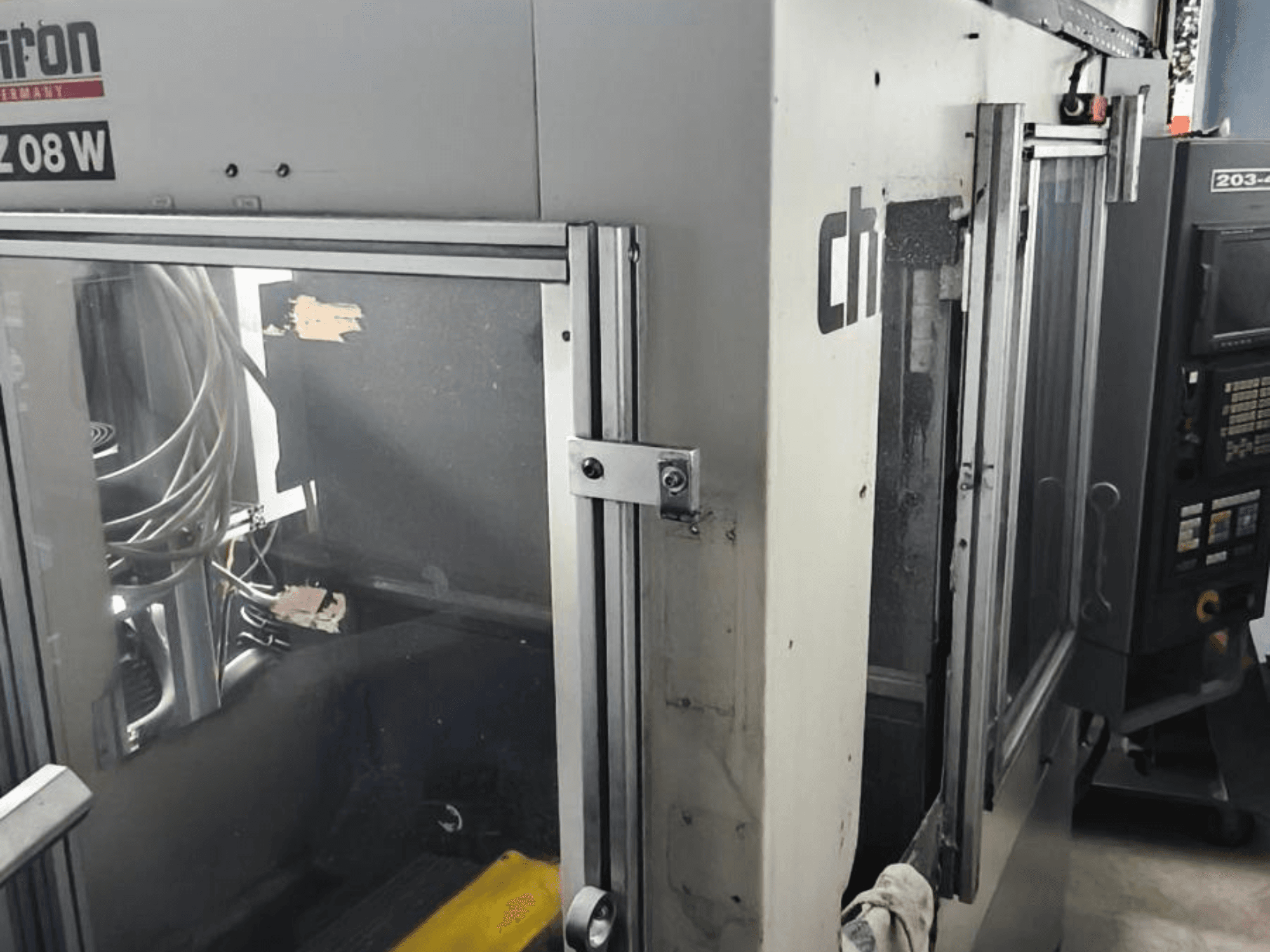 Vertikālais apstrādes centrs CHIRON FZ 08 W, priekšējais skats, kurā redzams vadības panelis un daļēji atvērtas durvis, ar redzamām iekšējām detaļām.