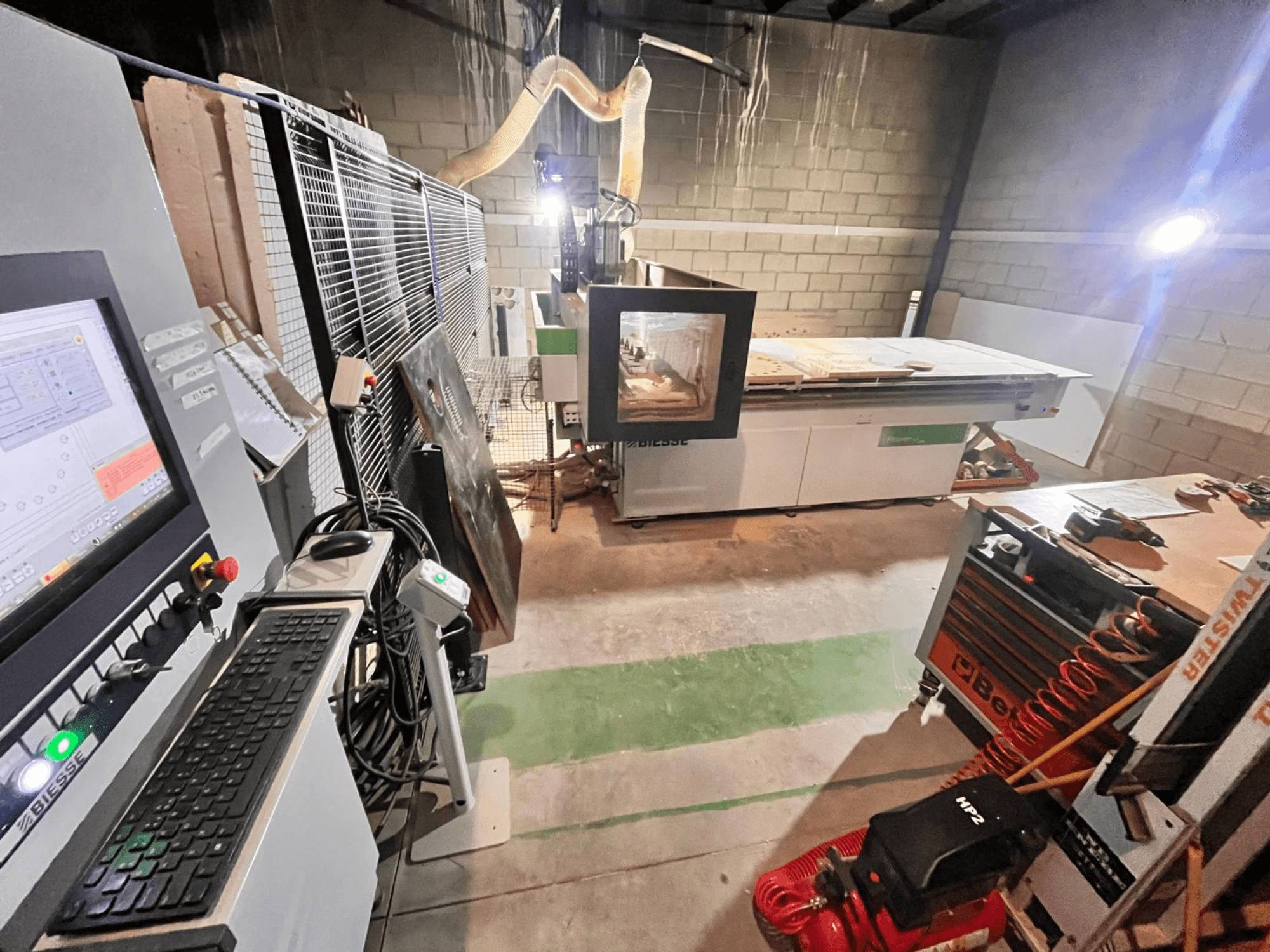 Biesse CNC darbnīcā, sānu skats, kurā redzams vadības panelis, putekļu nosūcējs, instrumenti un dažādi materiāli uz galda.