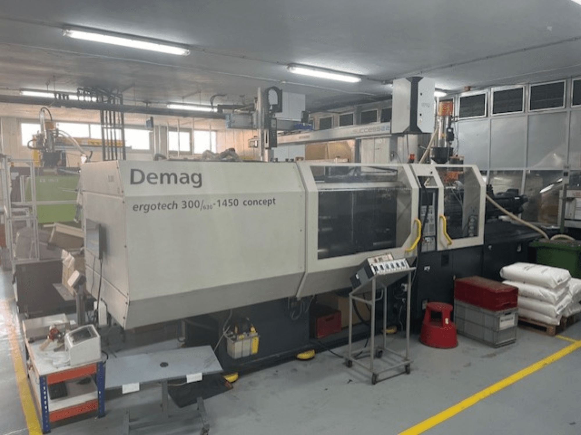 Demag ERGOTECH CONCEPT 3000-1450 hidrauliskā iesmidzināšanas iekārta, skats no priekšpuses, ar vadības ierīcēm un materiāliem blakus.