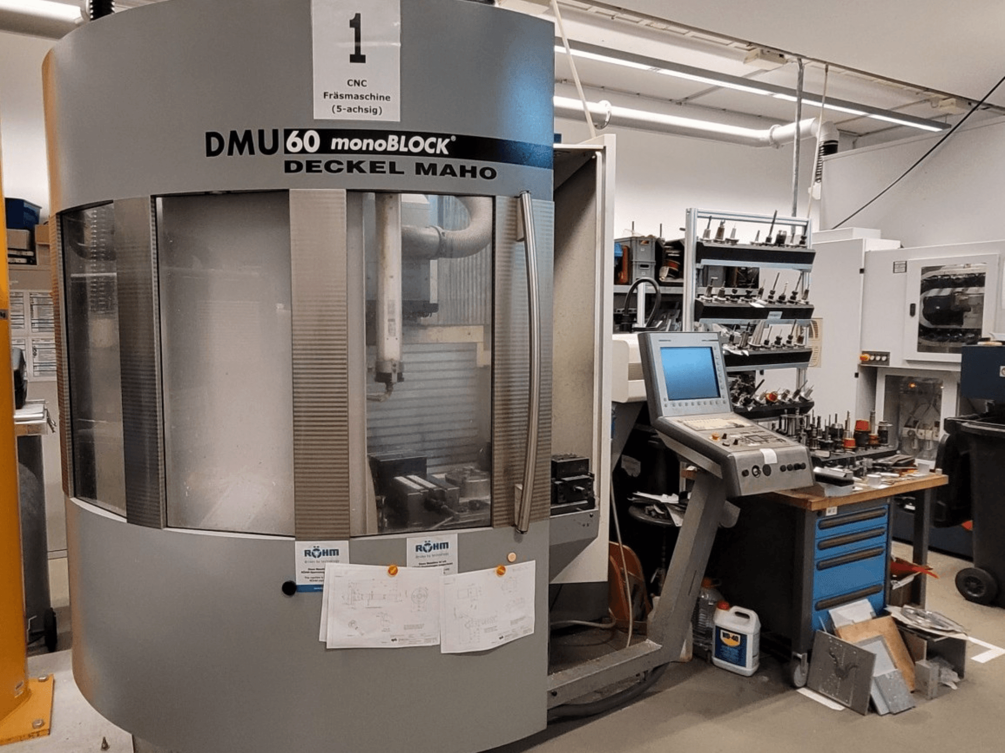 CNC frēzmašīna DECKEL MAHO DMU 60 monoBLOCK priekšpusē, blakus redzams vadības panelis un instrumentu noliktava.