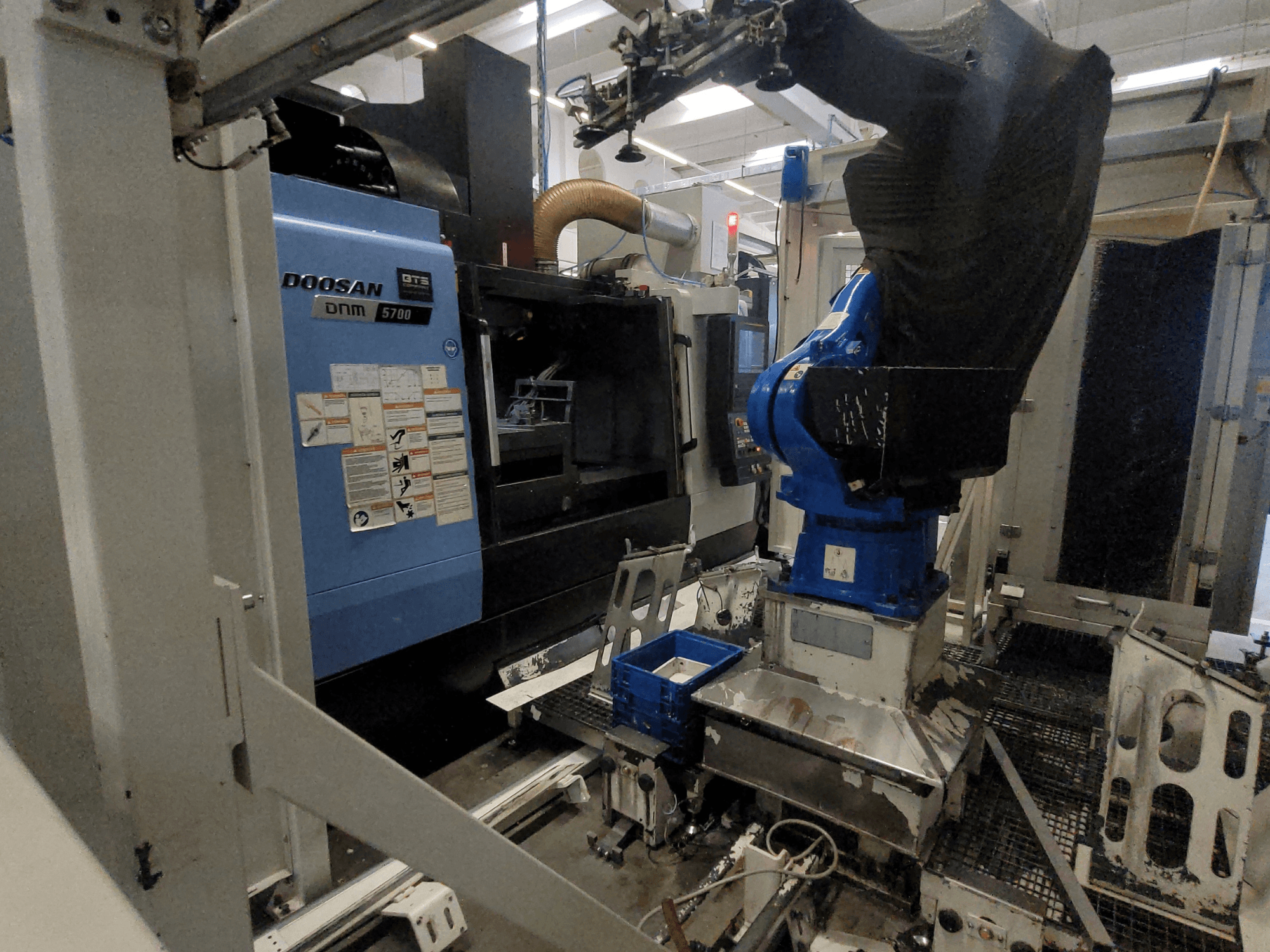 Vertikālais apstrādes centrs DOOSAN DNM 5700 rūpnīcas vidē, kurā redzams tā priekšskats un integrētā robota roka.