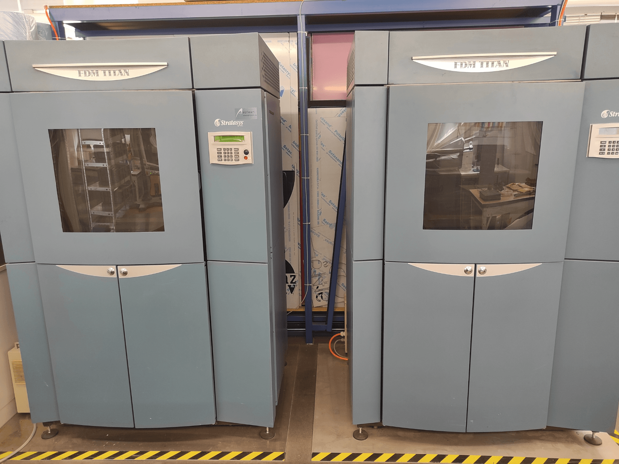 Stratasys ražotais FDM TitanTI 3D printeris, skats no priekšpuses, ar divām durvīm un vadības paneli, novietots rūpnieciskā vidē.