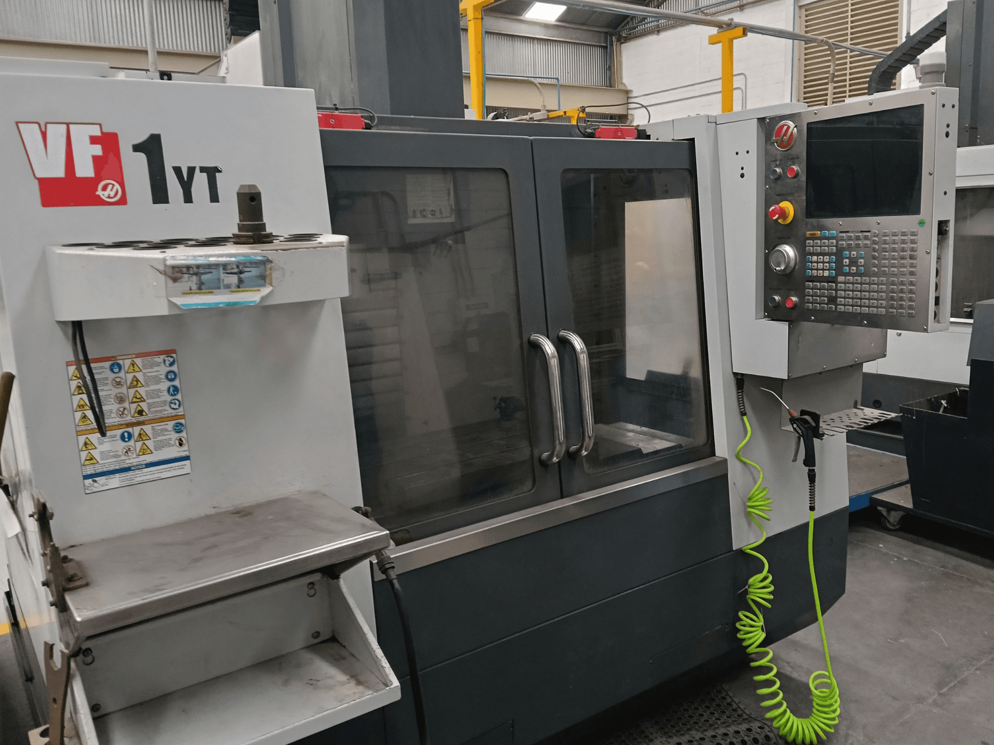 HAAS VF-1YT-2 vertikālais apstrādes centrs, skats no priekšpuses, kurā redzams vadības panelis, instrumentu turētājs, drošības uzlīmes un apstrādes zona.
